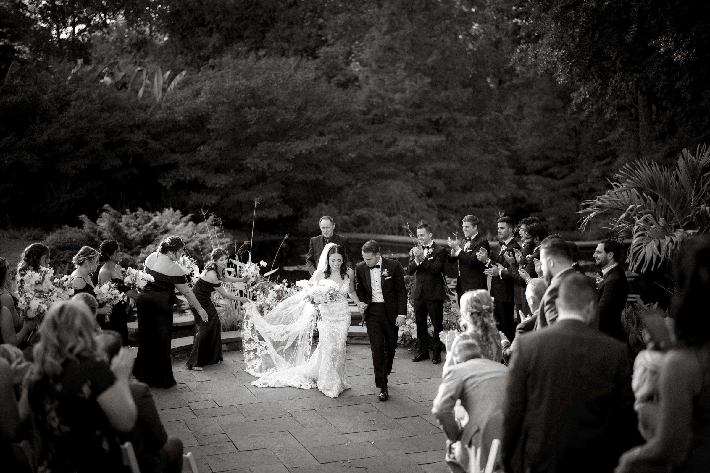 A bride and groom walk down the aisle at Duke Gardens, surrounded by cheering guests. The bride is in a flowing white dress, and the groom is in a suit. The outdoor setting features trees and lush foliage as guests clap and capture photos of this enchanting moment.