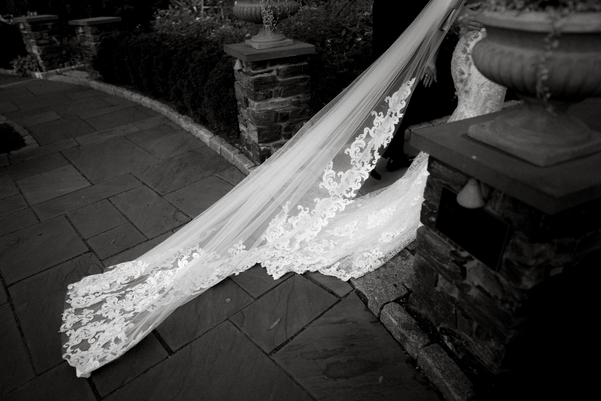 An elegant, lace-trimmed wedding veil flows gracefully onto a stone pathway beside a tiered stone urn at Duke Gardens. The lush greenery enhances the romantic atmosphere of this enchanting wedding setting.