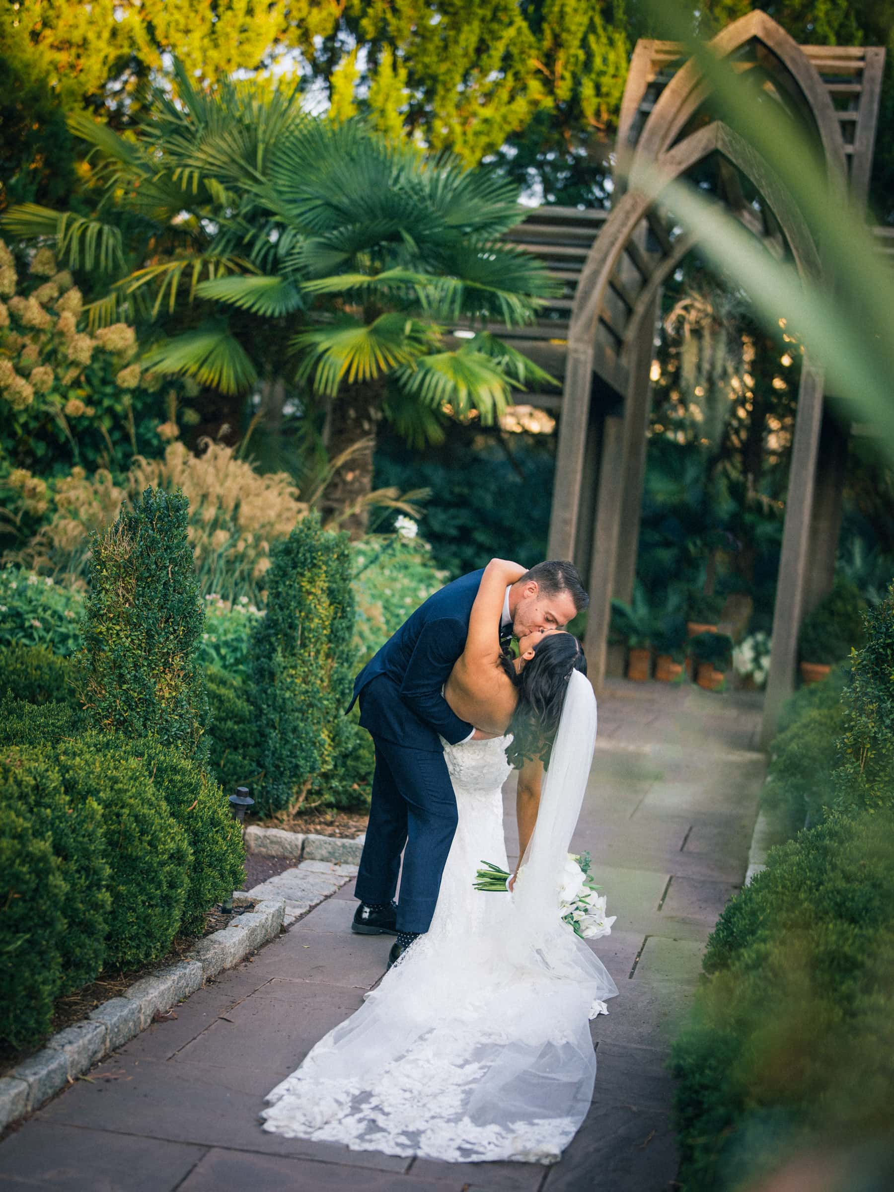A couple in wedding attire share a romantic kiss amidst the lush beauty of Duke Gardens. The bride, in a long white gown and veil, leans back as the groom holds her. They're surrounded by greenery and a charming wooden archway, capturing the essence of an enchanting garden wedding.