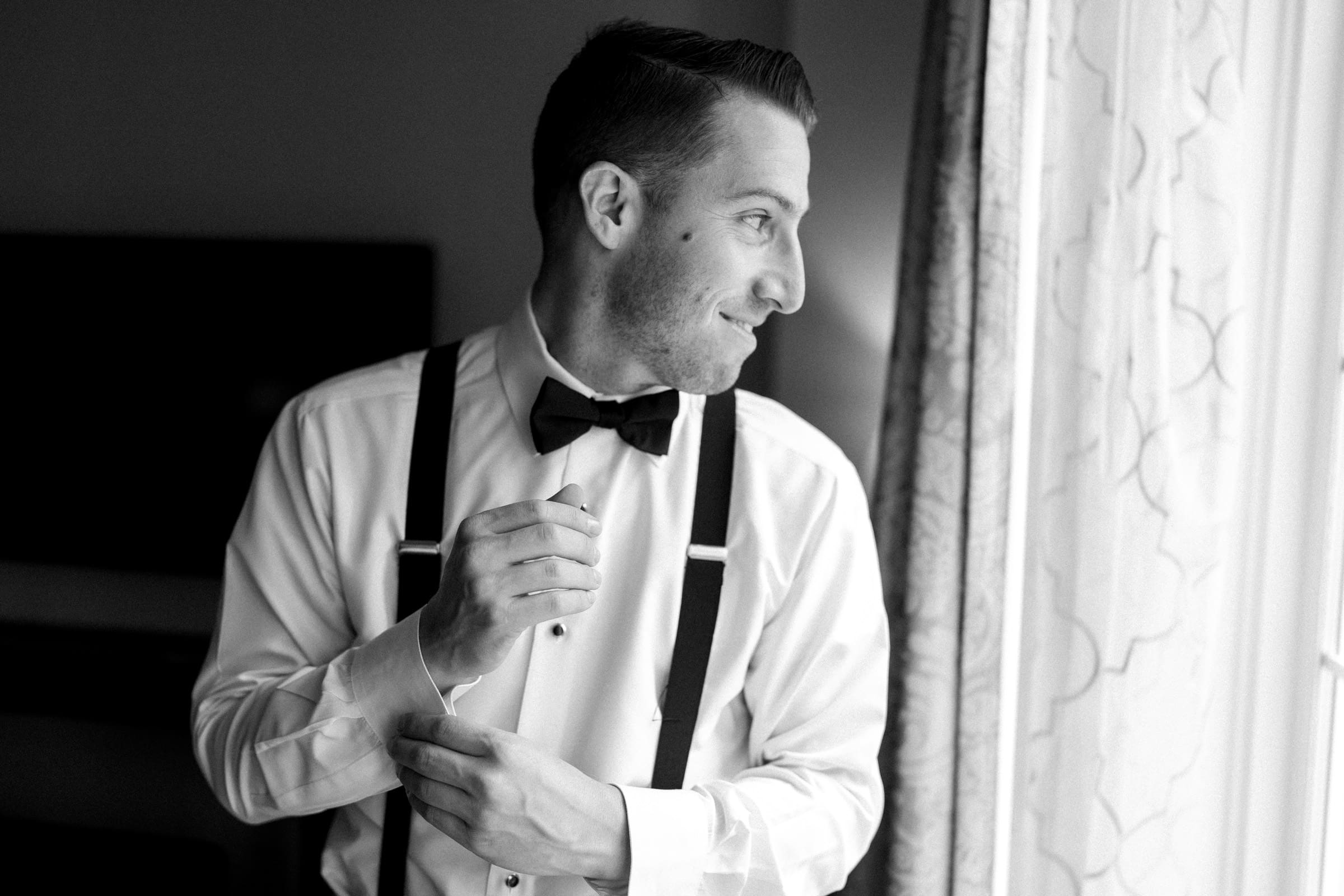 A man in a white shirt with suspenders and a bow tie smiles as he adjusts his outfit, envisioning the charm of a Duke Gardens wedding. He stands by the window, gazing outside. The timeless image is captured in black and white.