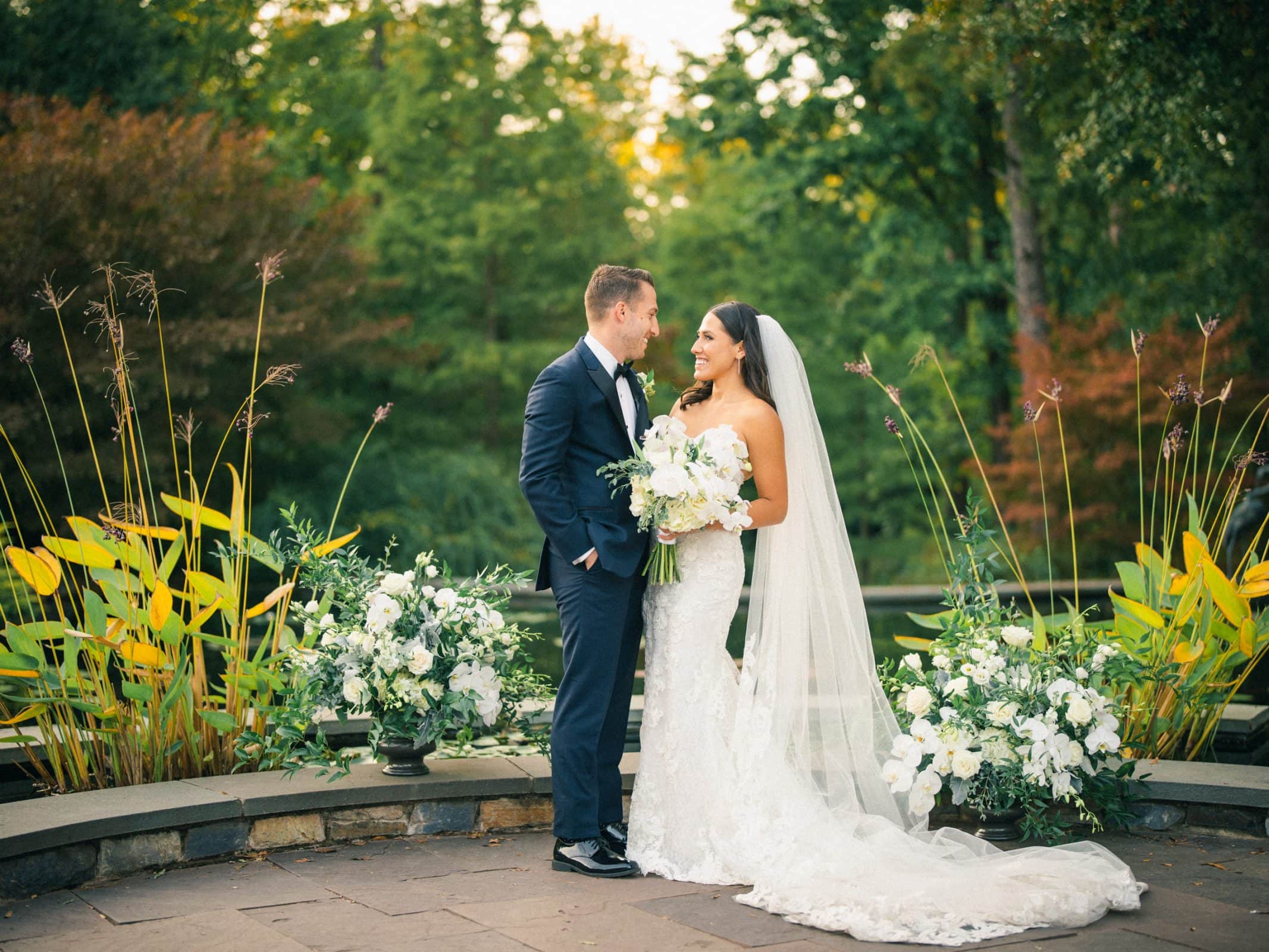 A couple stands in Duke Gardens, dressed in wedding attire. The bride, in a white gown and veil, holds a bouquet as they gaze at each other. The groom wears a dark suit. Lush greenery and white flowers surround them, creating a romantic setting for their Duke Gardens wedding.