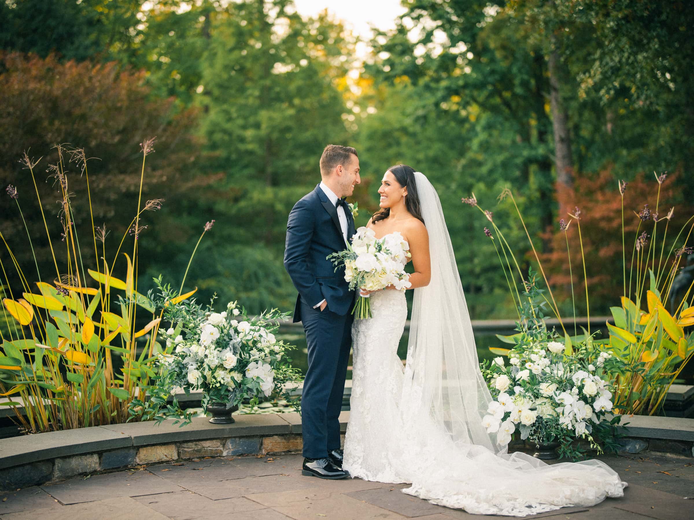 A couple stands in Duke Gardens, dressed in wedding attire. The bride, in a white gown and veil, holds a bouquet as they gaze at each other. The groom wears a dark suit. Lush greenery and white flowers surround them, creating a romantic setting for their Duke Gardens wedding.