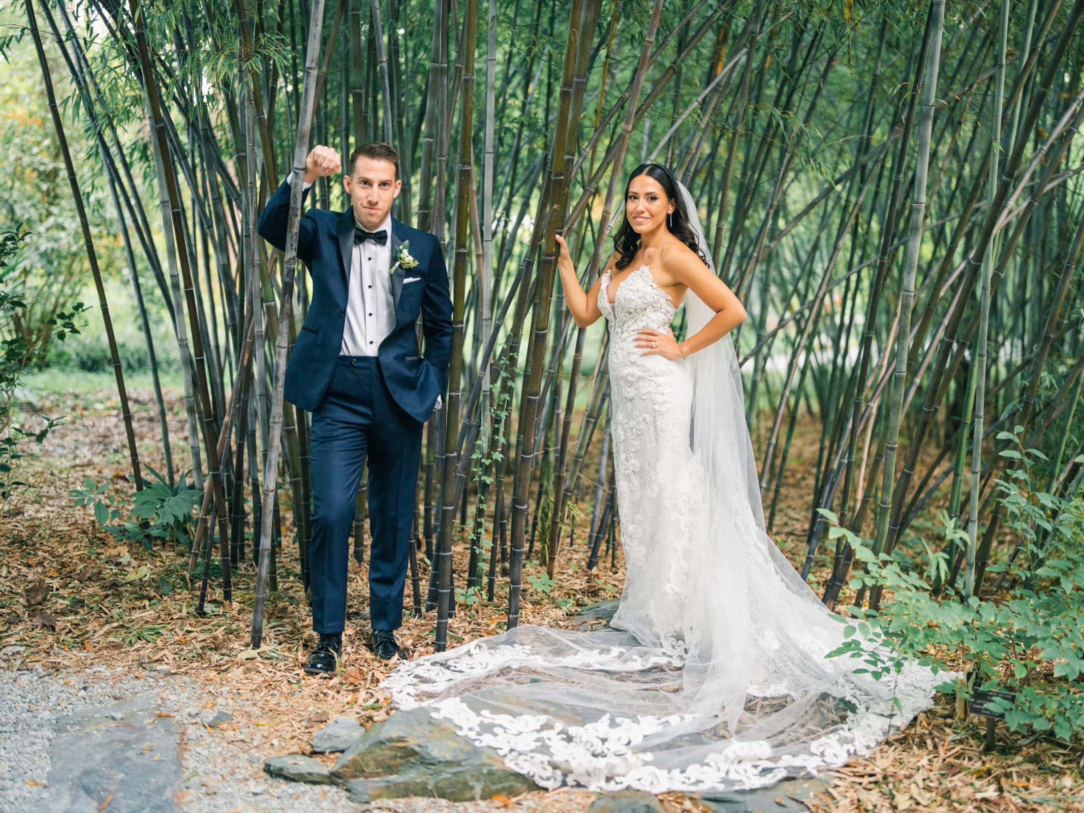A bride and groom pose in a bamboo forest during their Duke Gardens wedding. The groom, in a dark suit, casually holds his jacket over his shoulder, while the bride dazzles in a white lace dress with a long train. They stand on a path enveloped by lush greenery.