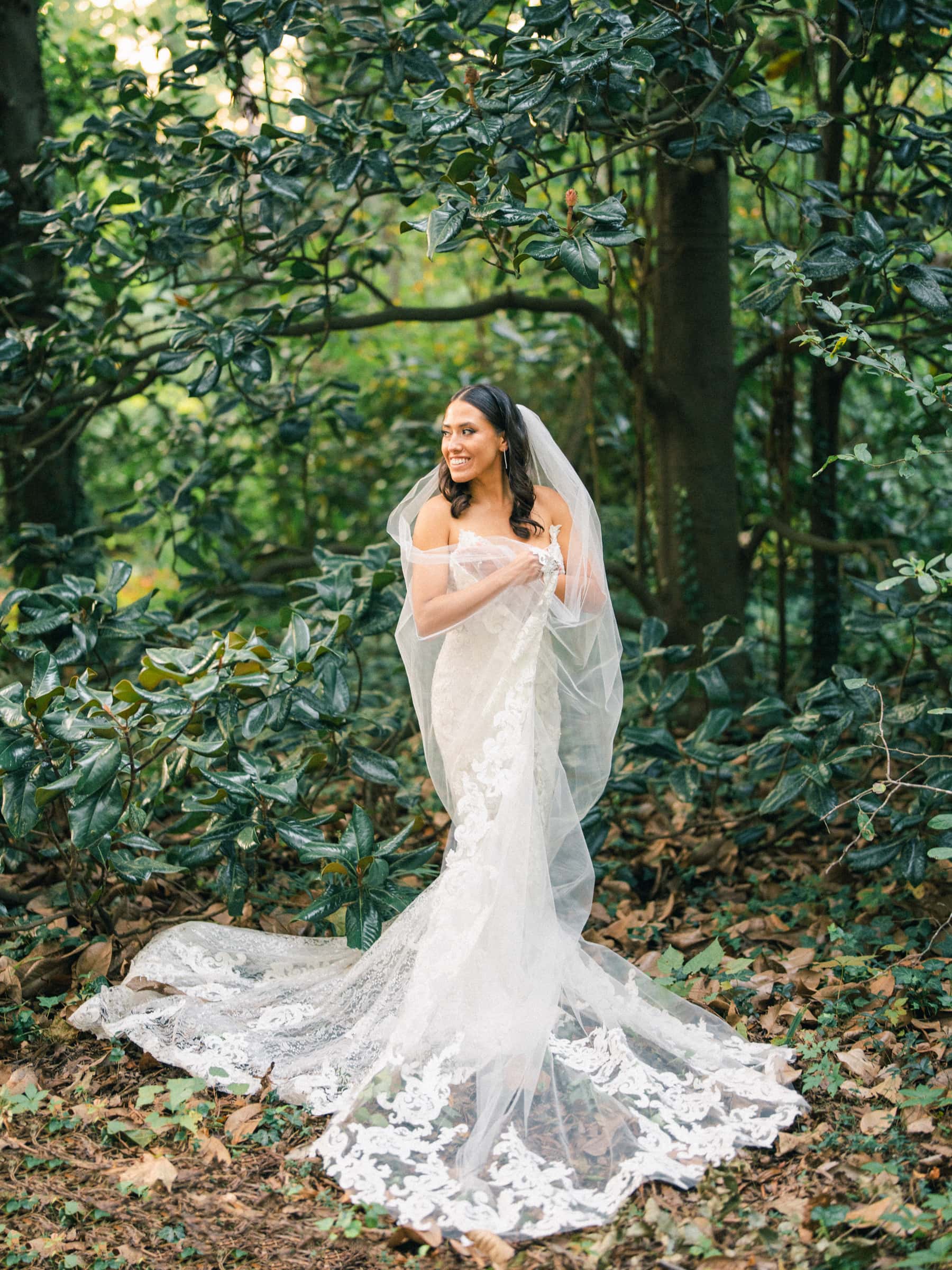 A bride stands in a forest at Duke Gardens, wearing a white lace wedding gown with a long train and veil. She is smiling and holding her veil with one hand, surrounded by lush greenery and fallen leaves.