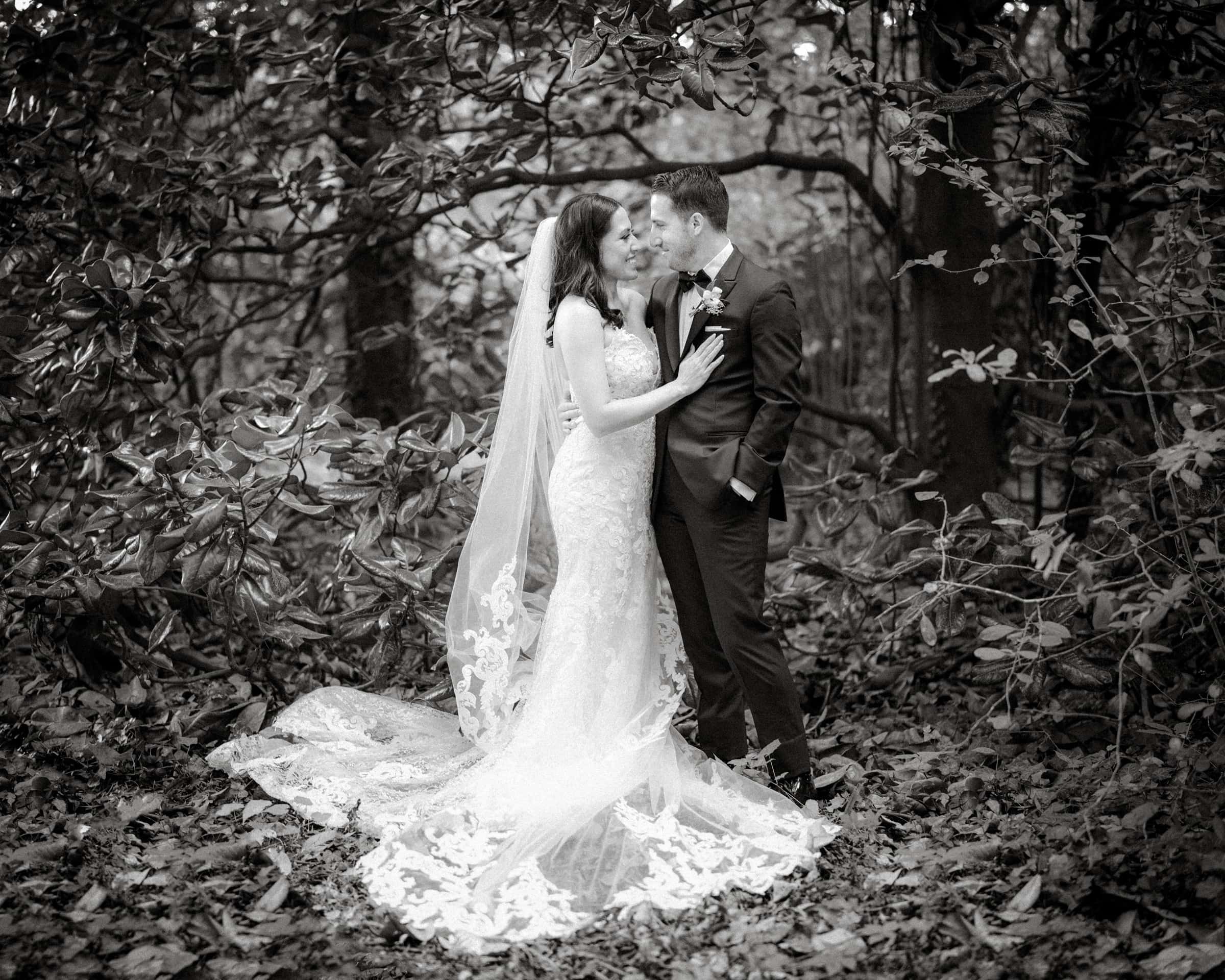 A bride in a lace gown and a groom in a suit stand close, smiling at each other beneath the canopy of Duke Gardens. The bride wears a long veil that flows onto the leaves, adding to the romantic, nature-filled setting.