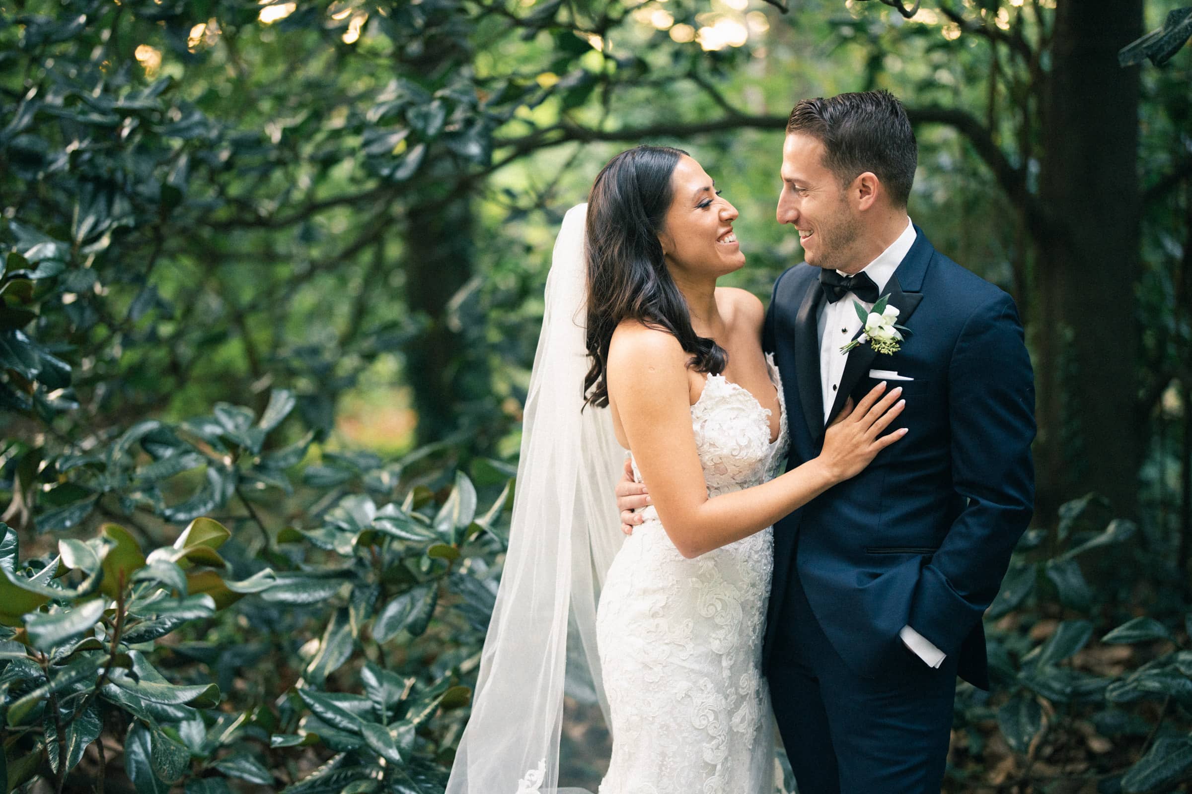 A bride and groom stand in a lush, green garden at Duke Gardens. The bride wears a lace wedding dress and veil, smiling at the groom in a dark suit and bow tie. They are surrounded by dense foliage, creating a romantic atmosphere perfect for their wedding day.