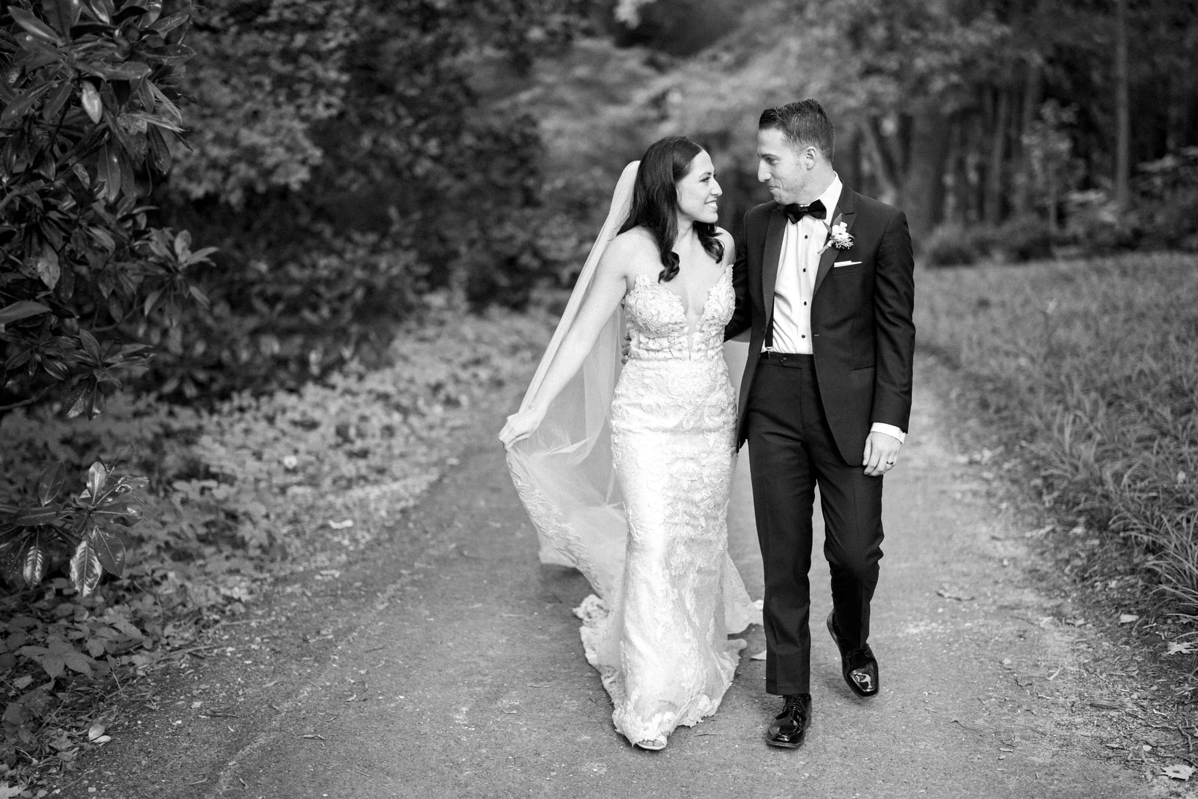 A bride and groom walk hand in hand down a tree-lined path at Duke Gardens. The bride wears a lace wedding dress with a veil, and the groom is in a tuxedo. Both are smiling as they look at each other. The nostalgic scene is captured in black and white.