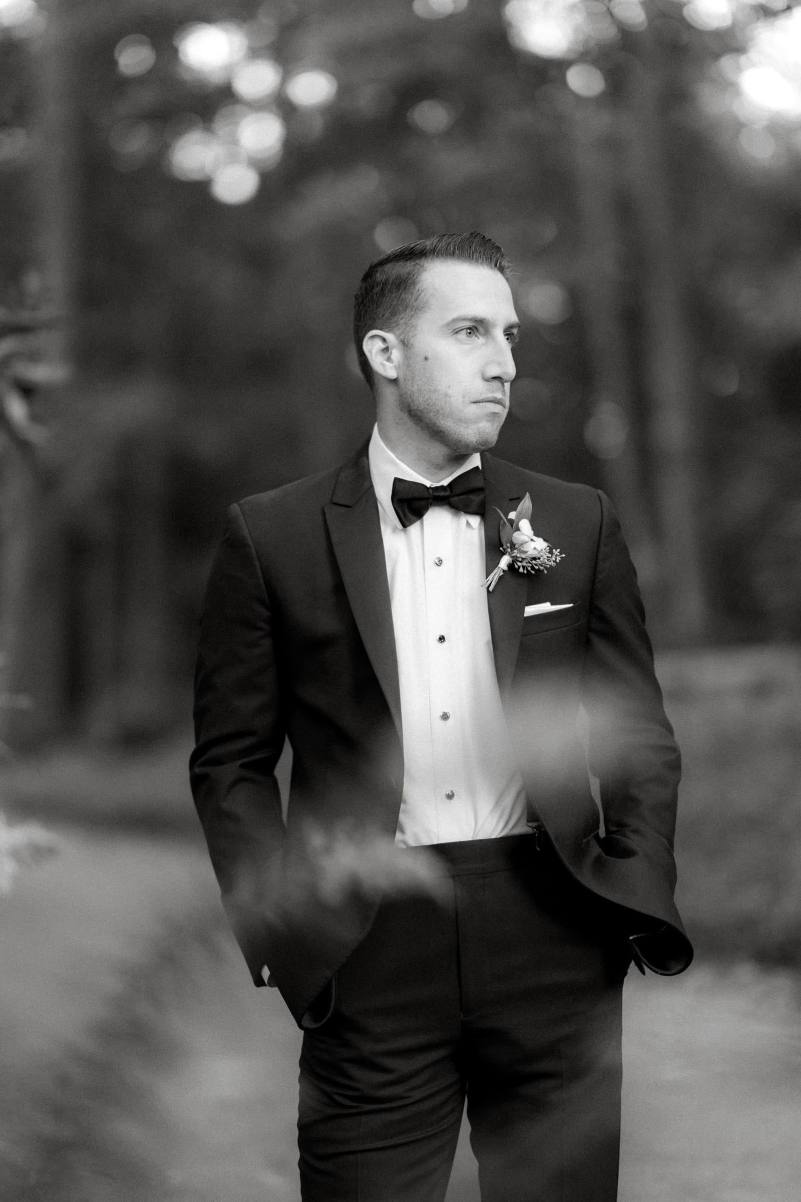 A black and white photo captures a man in a suit and bow tie standing outdoors at Duke Gardens. With his hands in his pockets, he gazes off to the side, the softly blurred background highlighting the enchanting forest setting of a wedding day.