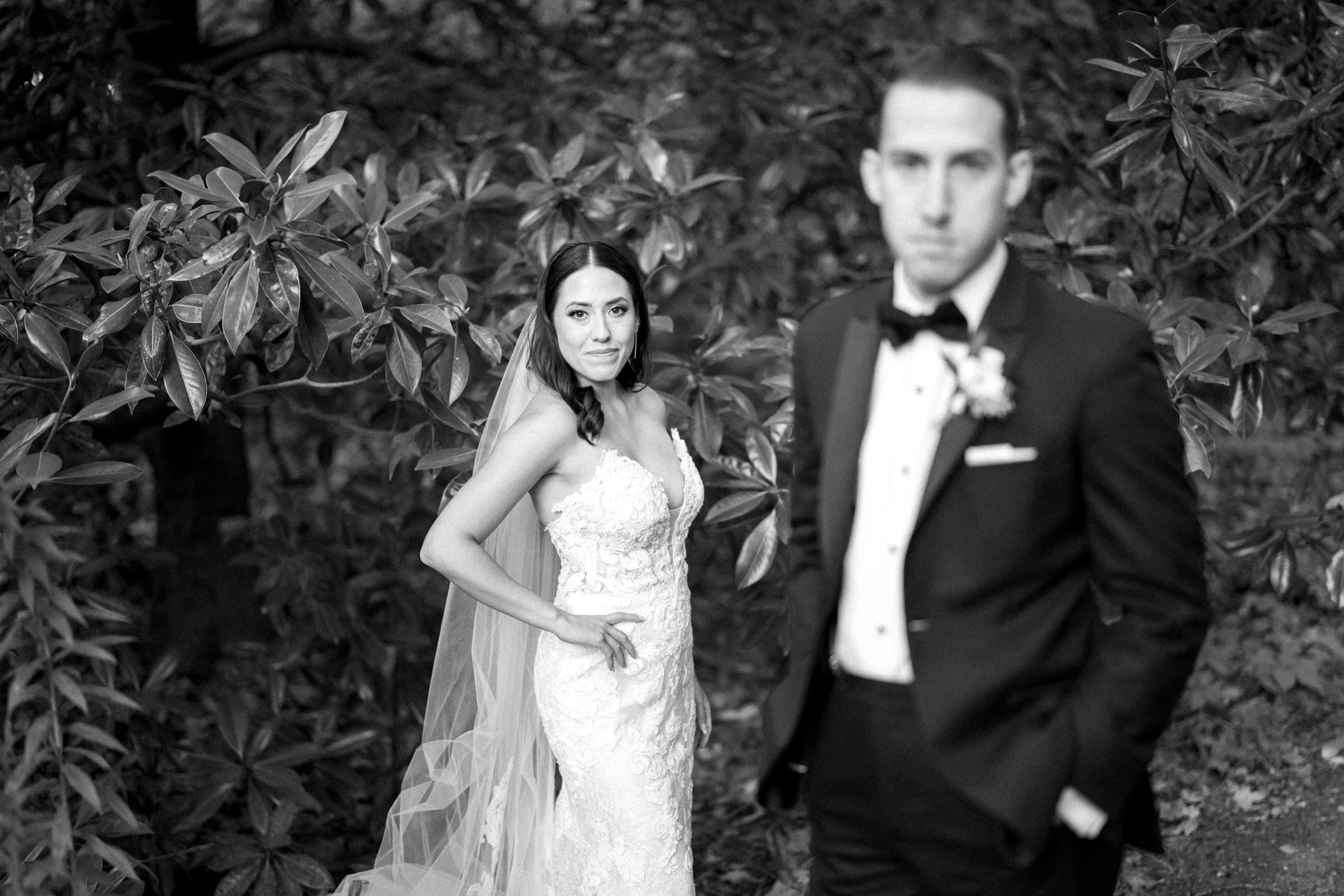 A bride in a lacy wedding dress stands confidently with one hand on her hip, looking at the camera. A groom in a tuxedo, set against the lush greenery of Duke Gardens, stands slightly blurred in focus, adding to the enchanting ambiance of their wedding day.