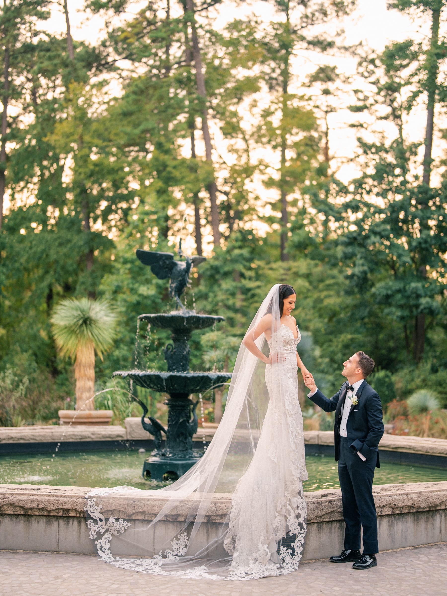 A bride in a long veil and lace dress stands on a ledge, holding hands with her groom in a tuxedo. They are posed in front of the large, ornate fountain at Duke Gardens, with tall trees providing a lush backdrop to their enchanting wedding scene.