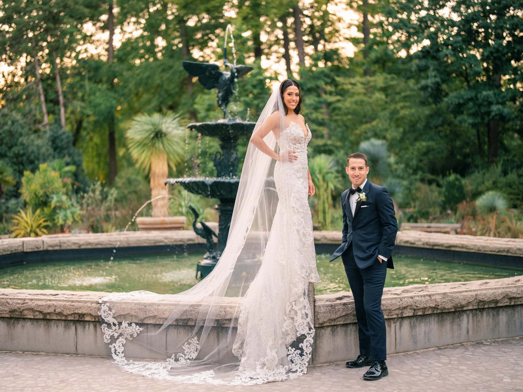 A bride in a lace wedding gown and long veil stands on the edge of a fountain next to her groom in a dark suit and bow tie. Surrounded by lush greenery, she smiles radiantly while he looks at the camera, perfectly capturing the enchanting charm of this Duke Gardens wedding.