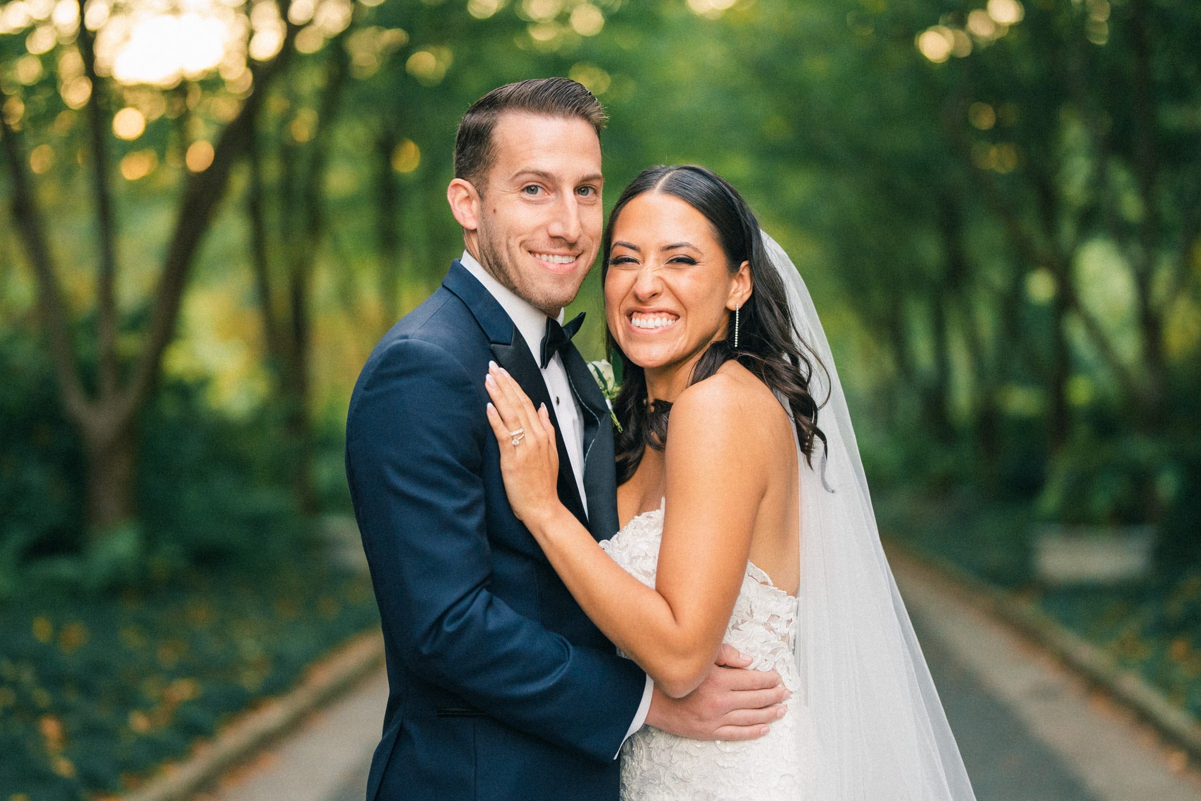 A bride and groom smile joyfully at the camera in an outdoor setting, capturing the essence of a Duke Gardens wedding. The bride, wearing a white dress and veil, embraces the groom in a dark suit as they stand on a tree-lined path with soft, natural lighting.
