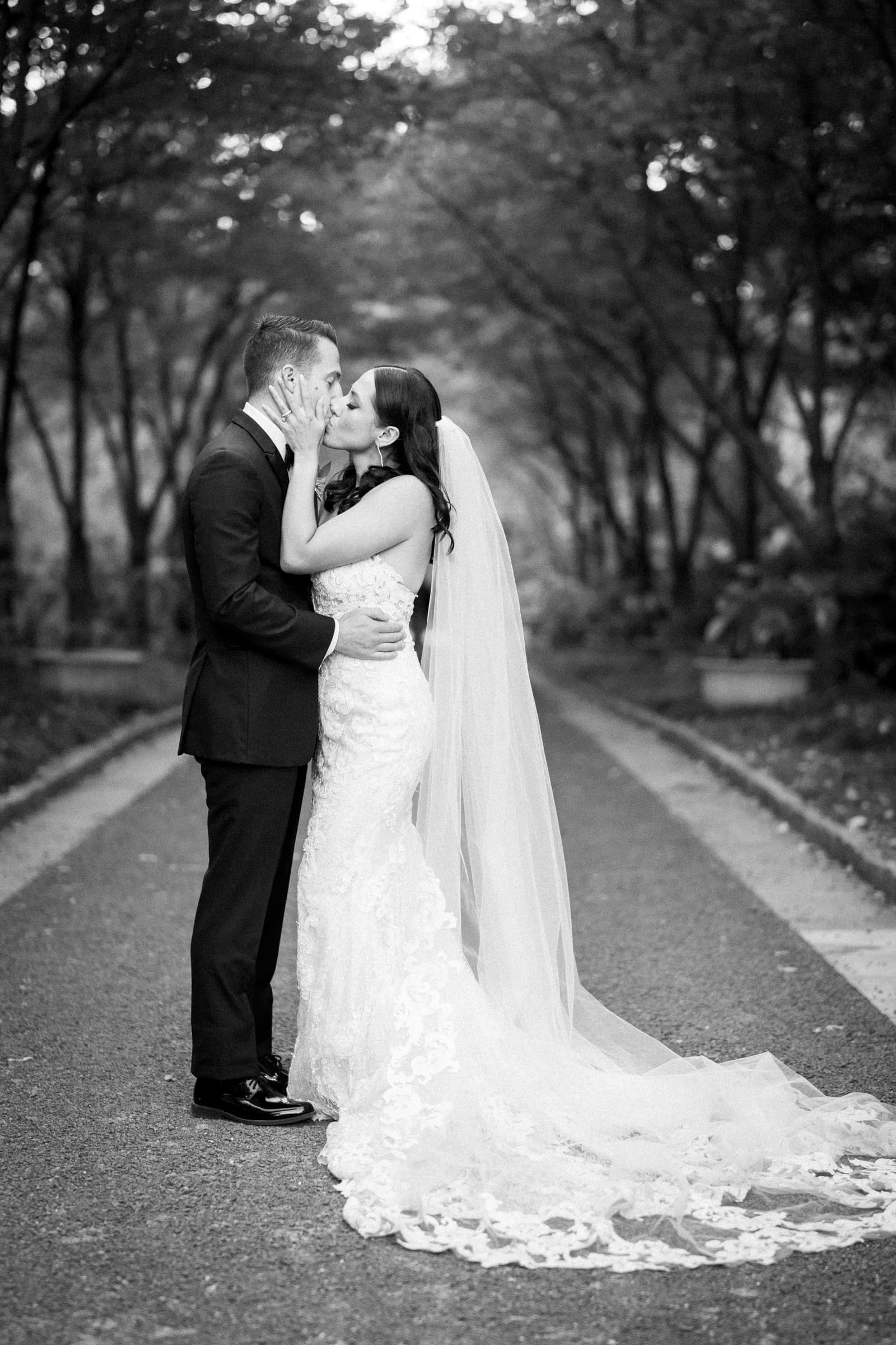 A bride and groom kiss on a tree-lined path at Duke Gardens. The bride wears a long lace dress and veil, while the groom is in a dark suit. This black-and-white photo beautifully captures their romantic outdoor wedding moment.