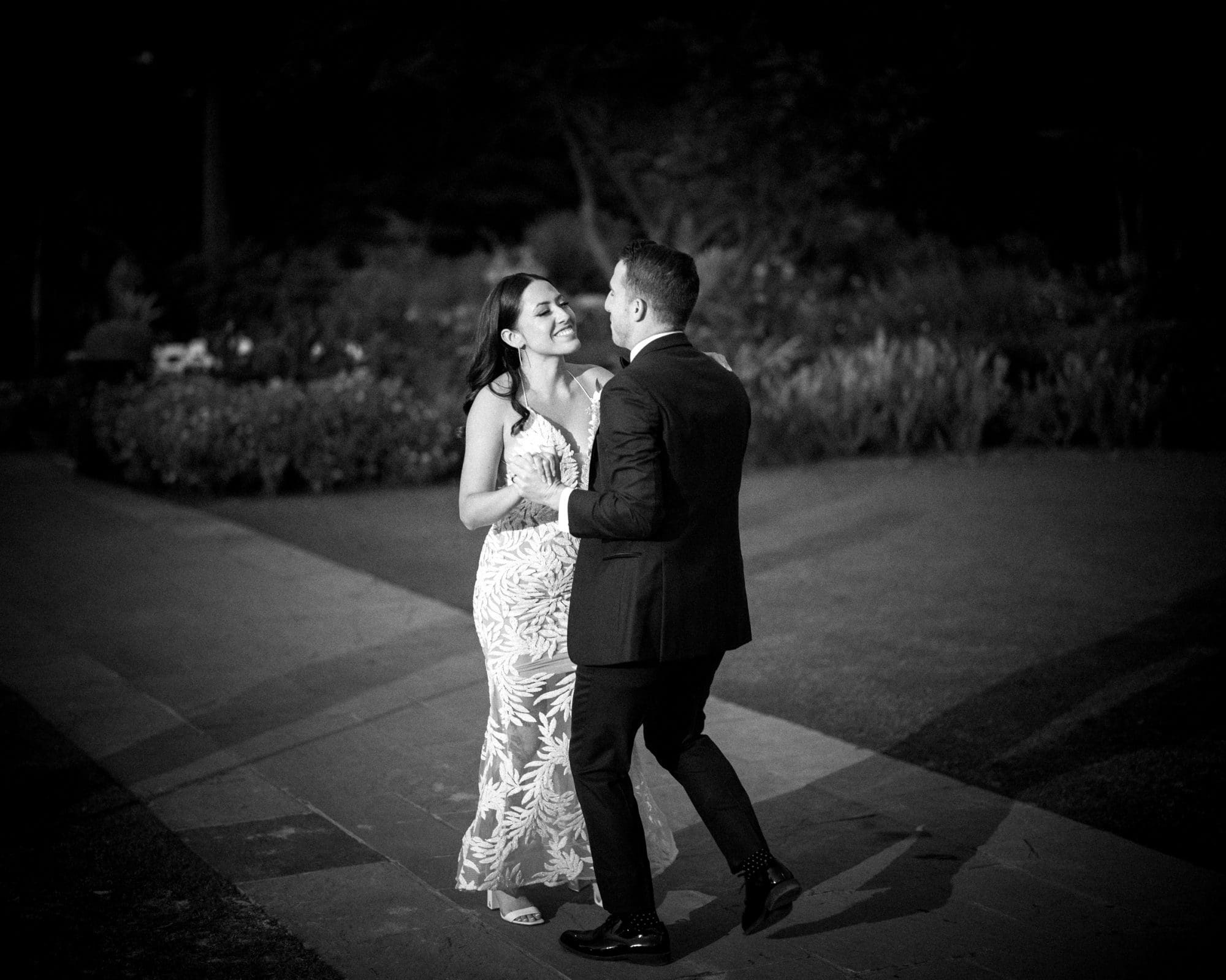 A couple dances together on a garden path at Duke Gardens, their tender moment evoking the elegance of a wedding night. The woman wears a patterned dress, and the man is in a suit. The scene is captured in black and white, with surrounding greenery in soft focus.