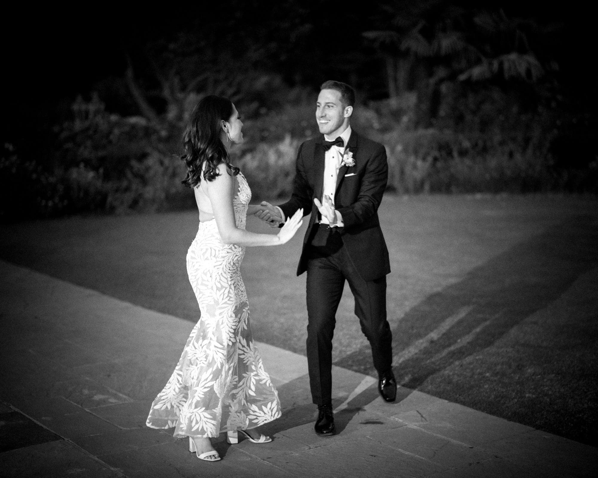 A black and white image captures a couple dancing outdoors at their Duke Gardens wedding. The man, in a tuxedo, reaches out to the woman in a floral dress. They are smiling and holding hands, with the lush garden serving as their enchanting backdrop.