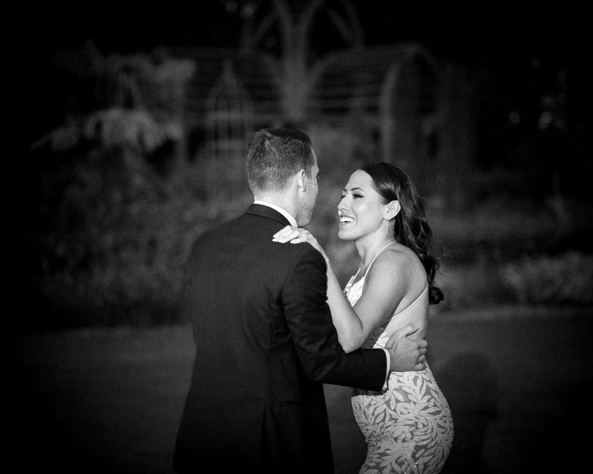 A couple dances joyfully in Duke Gardens, their figures framed by the timeless, black-and-white ambiance. The woman smiles in a patterned dress while the man, dapper in his suit, twirls her against softly blurred backgrounds—a perfect moment in this enchanting wedding setting.