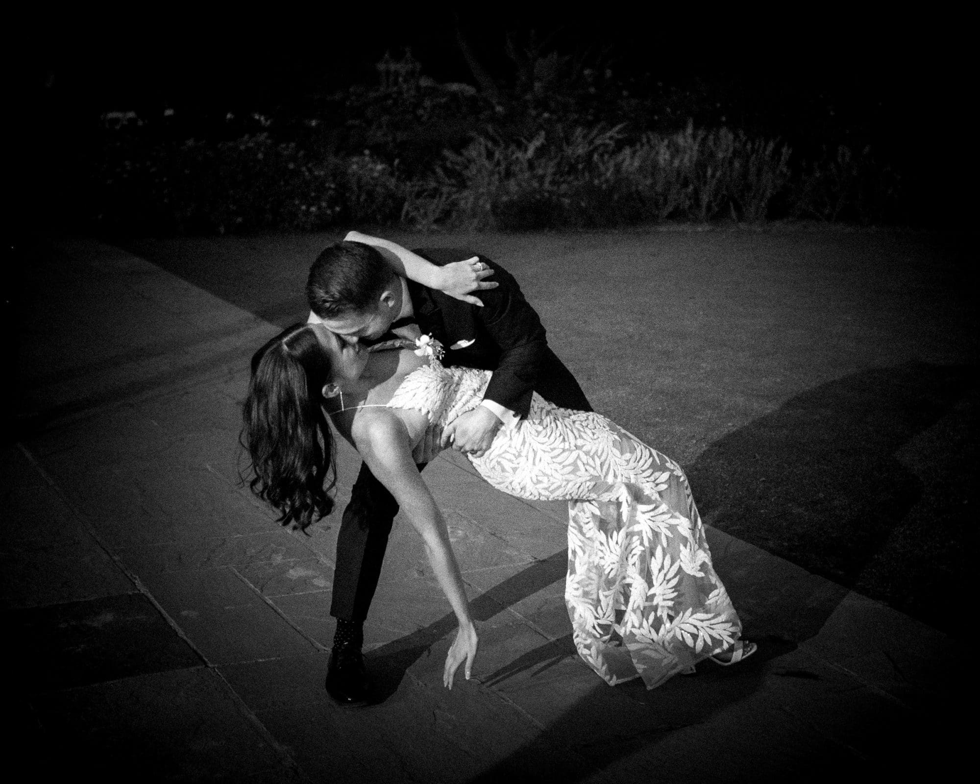 A couple dances under the night sky at Duke Gardens. The man in a suit dips the woman in a patterned dress as they kiss, capturing a romantic and elegantly lit moment with the lush garden providing a perfect wedding backdrop.