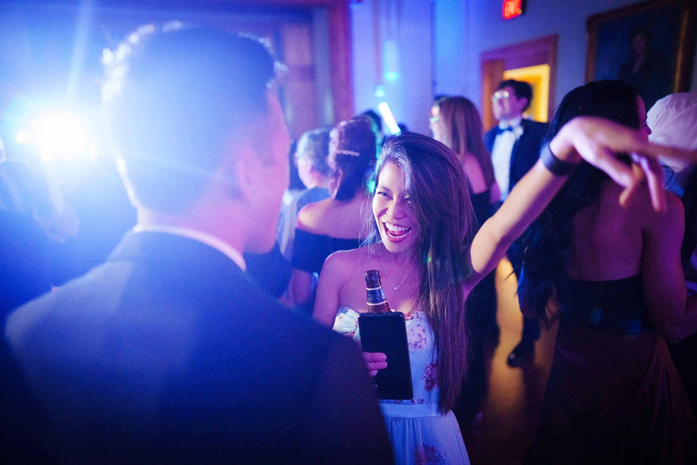 A lively party scene reminiscent of a Duke Gardens wedding, with guests in formal attire. A woman in the foreground is smiling and holding a drink, while others dance and socialize under colorful lights illuminating the room.