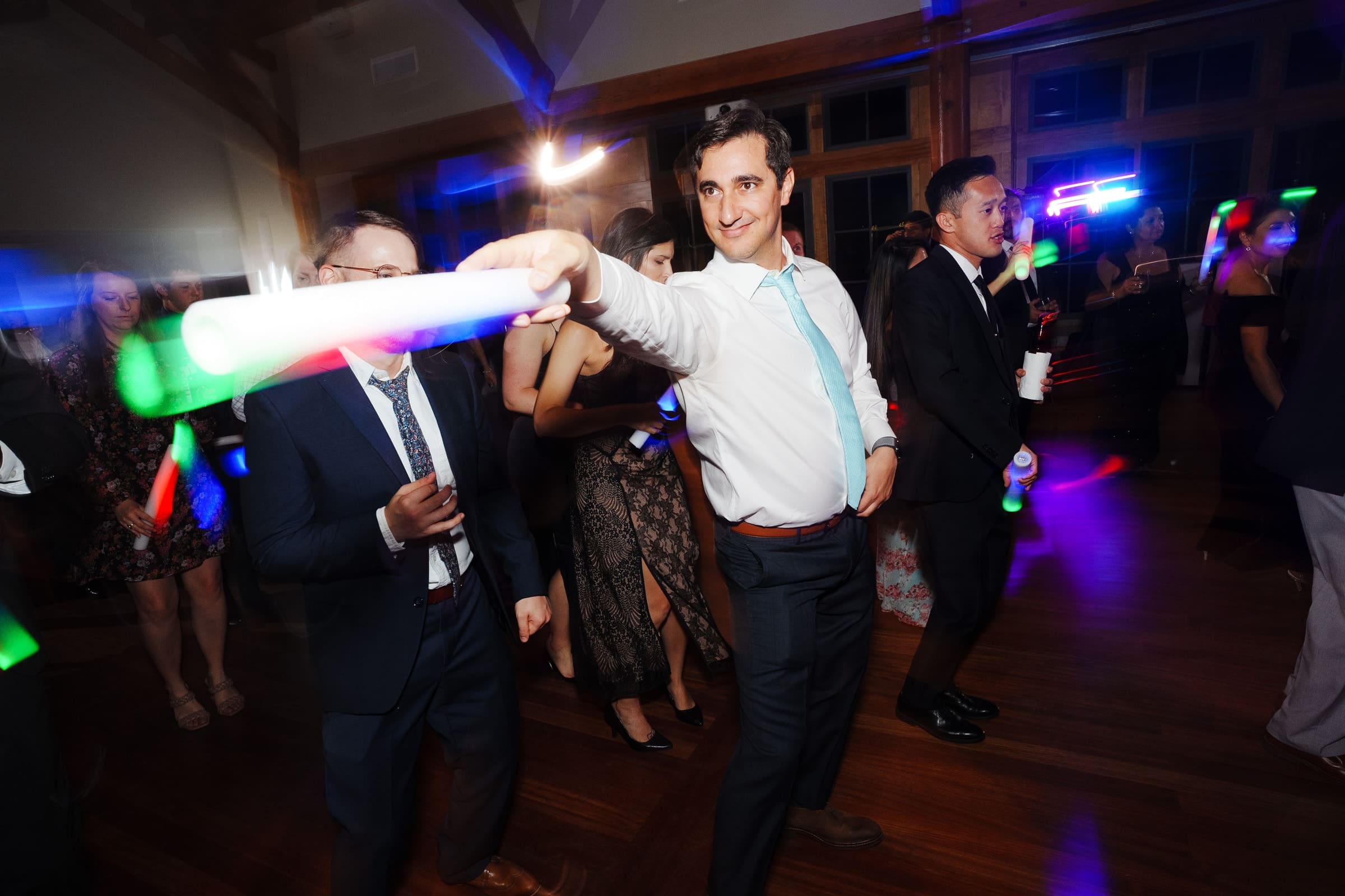 A man in a white shirt and tie dances energetically while holding a lit wand at a lively Duke Gardens wedding. Colorful lights and guests in formal attire fill the room, creating a festive atmosphere.