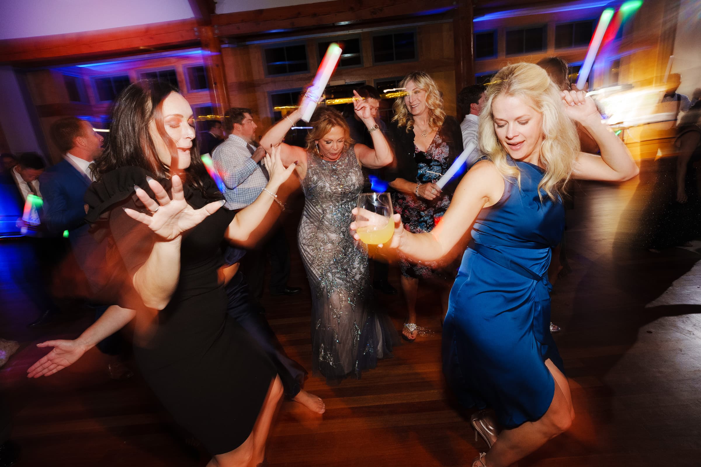 A lively dance floor scene unfolds at a Duke Gardens wedding, with guests reveling in the joyous celebration. Several women dance energetically, holding drinks and glow sticks, as the room is warmly lit with a festive ambiance that captures the magic of the evening.