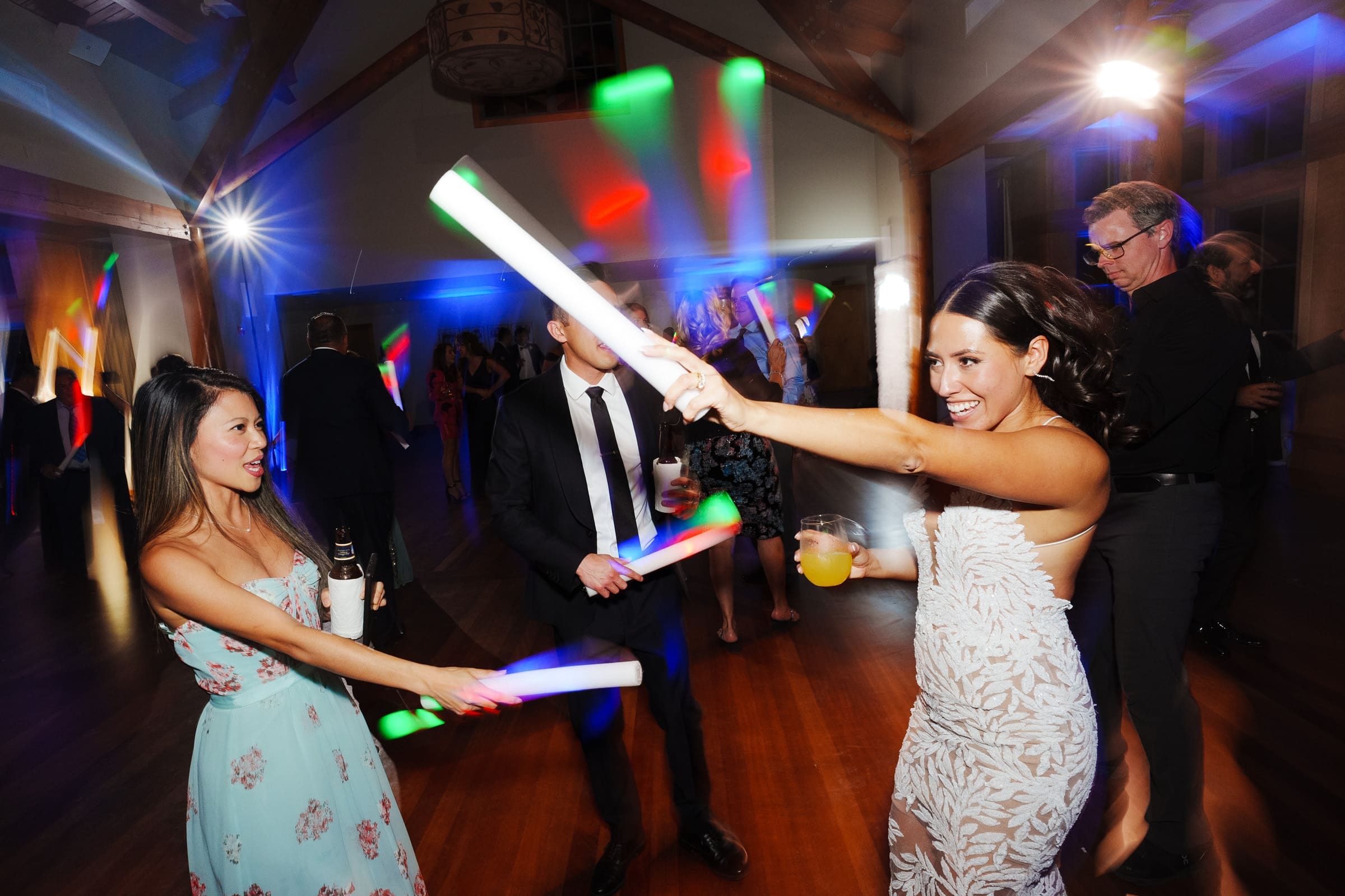 People dancing energetically at a party, holding glowing light sticks. The room, reminiscent of a Duke Gardens wedding, is filled with colorful lights and guests dressed in formal attire, enjoying the festive atmosphere.