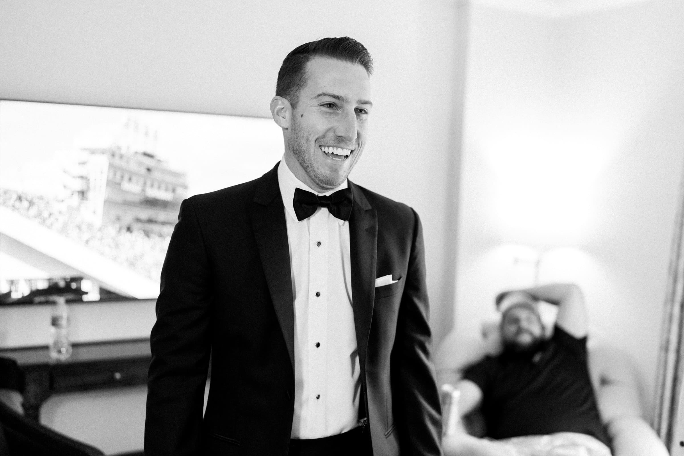 A man in a tuxedo smiles in a room, standing in front of a TV showing a sports scene, as if he's just returned from a Duke Gardens wedding. Another person sits in the background on a chair, wearing casual clothes and leaning back with a relaxed posture.