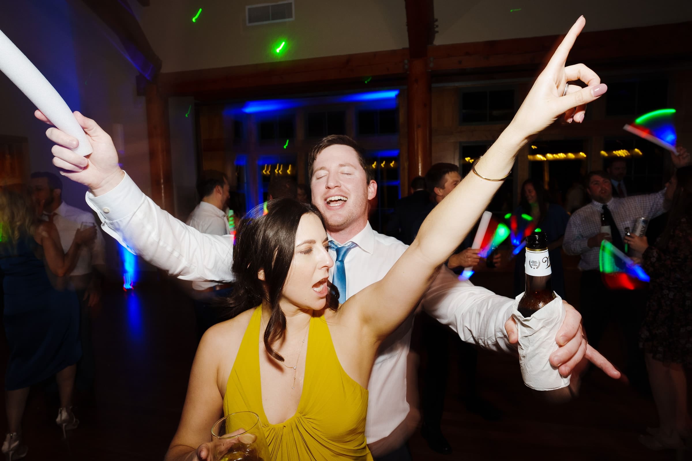 At a lively Duke Gardens wedding, a man and woman dance energetically. The woman in a yellow dress holds a drink, while the man in a tie raises his beer triumphantly. Colorful lights illuminate the festive crowd dancing joyfully in the background.