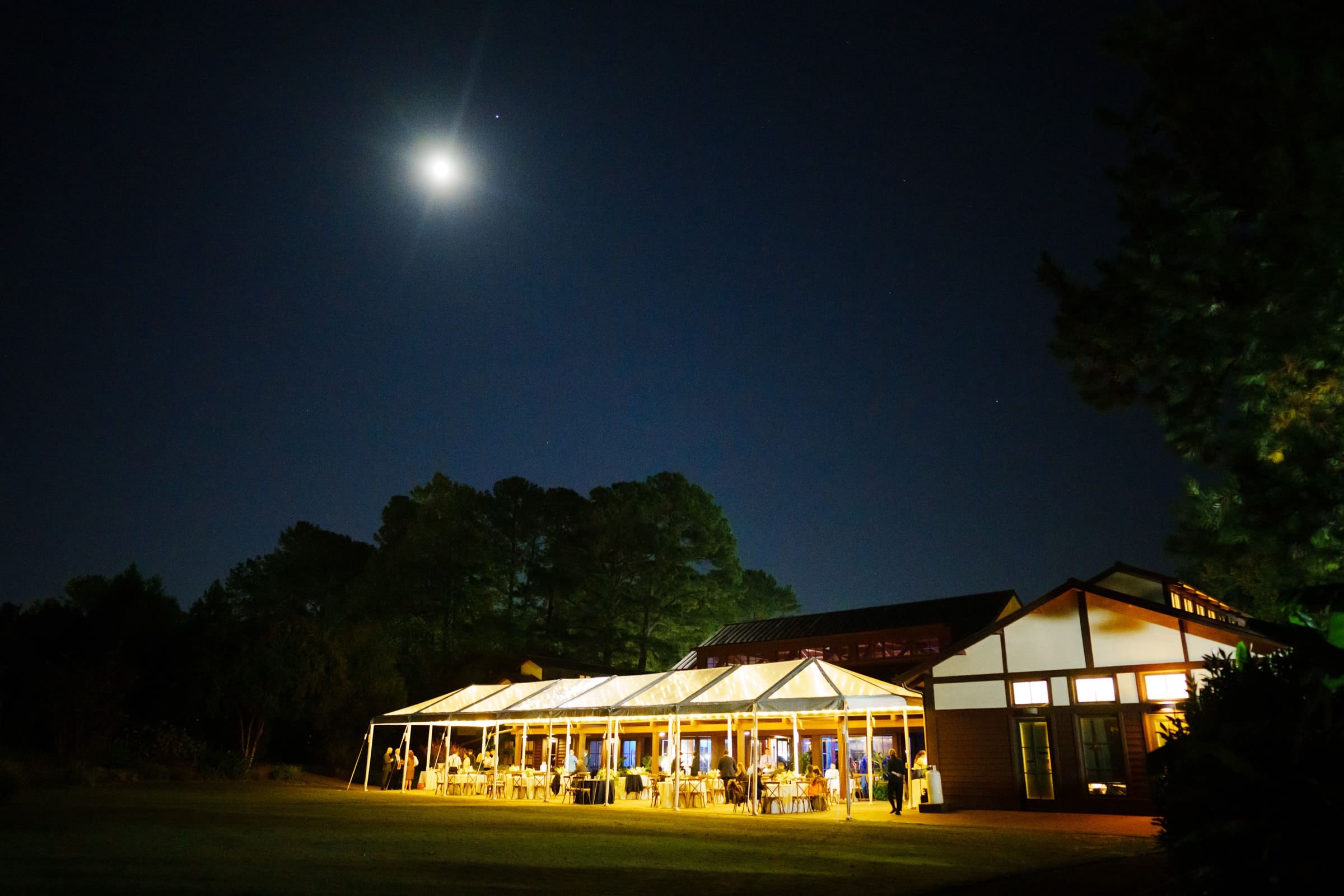 A nighttime wedding reception under a clear sky in Duke Gardens with a bright full moon. Guests are seated inside a well-lit, transparent tent adjacent to a large building, surrounded by trees.