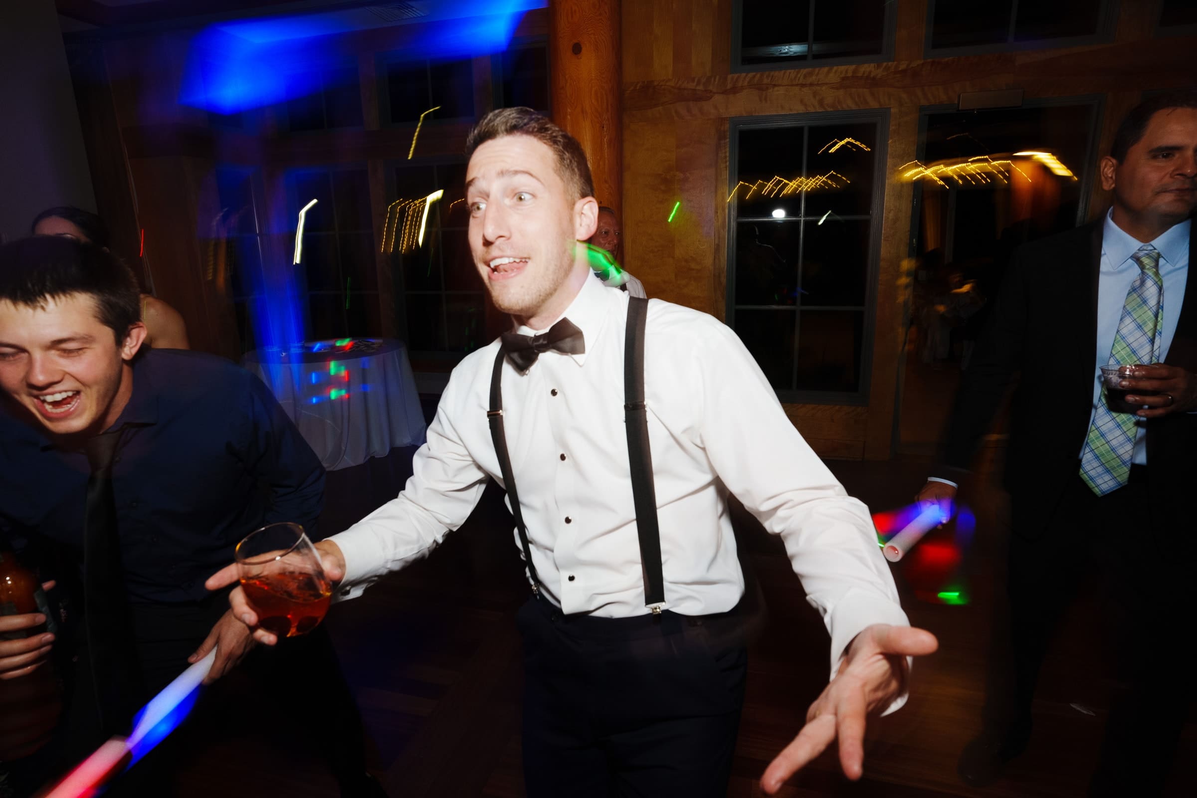 At a vibrant Duke Gardens wedding, a man in a tuxedo and suspenders dances energetically amid colorful lights. Holding a drink, he laughs along with others, as cheerful guests revel in the joyous atmosphere beside him.