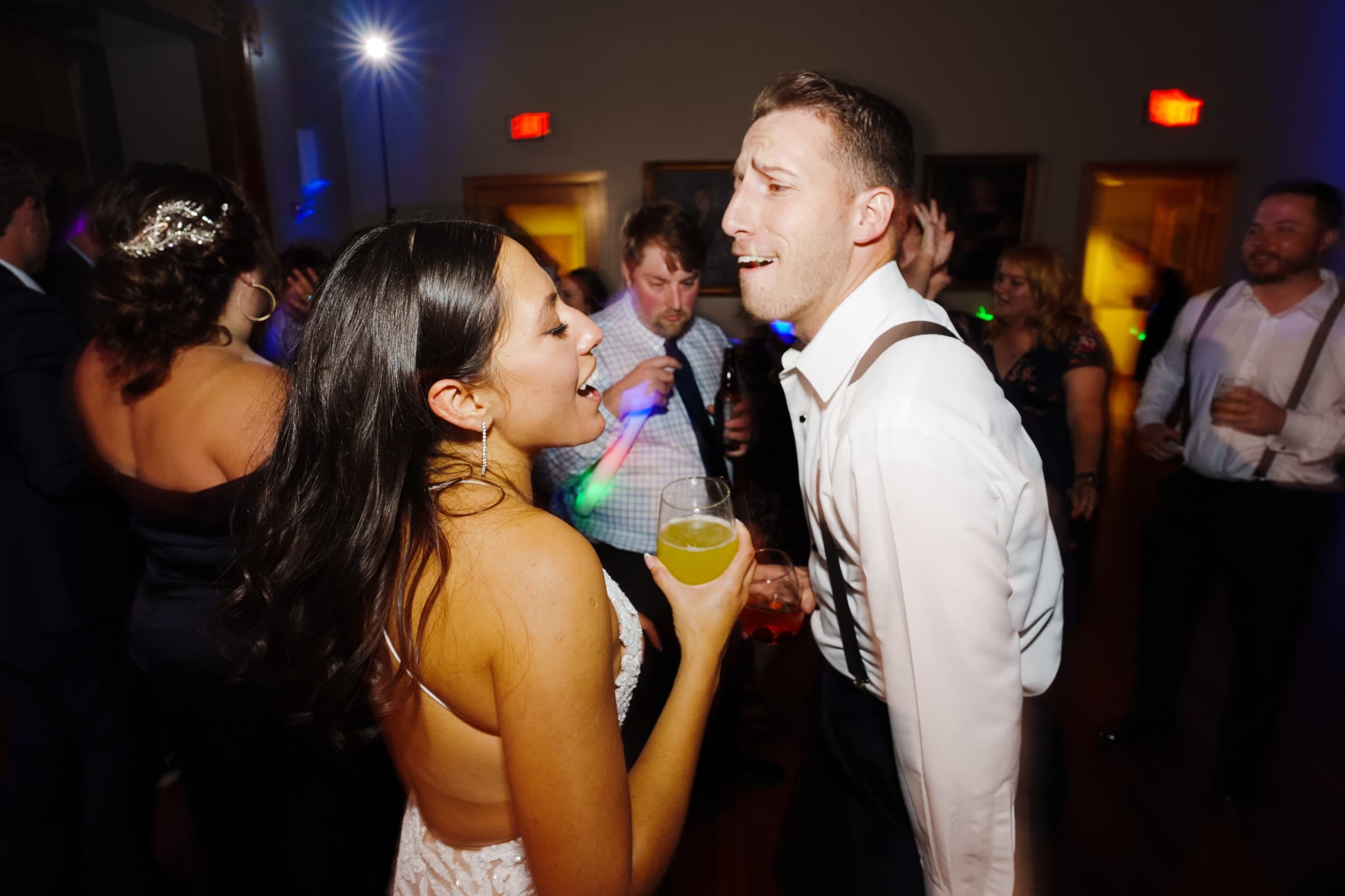 A couple joyfully singing and dancing together at a lively indoor party, reminiscent of a Duke Gardens wedding. The man holds a red drink, while the woman has a yellow beverage. Other guests, blurred in the background, are also enjoying the celebration.