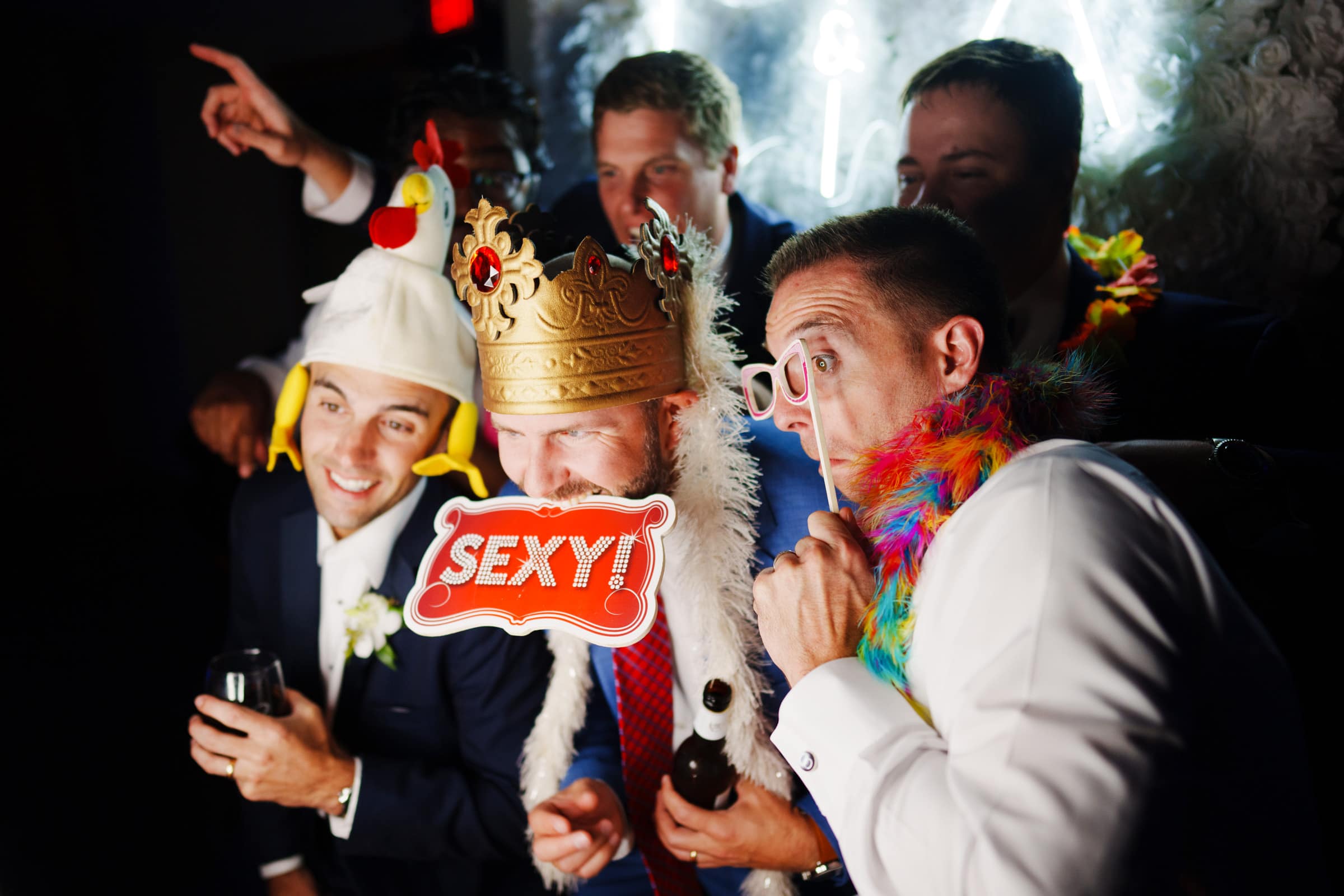A group of men at a party, wearing playful costumes including a chicken hat, crown, and feather boa. One holds a sign that says SEXY! They are smiling and posing together in front of a bright backdrop reminiscent of Duke Gardens wedding festivities.