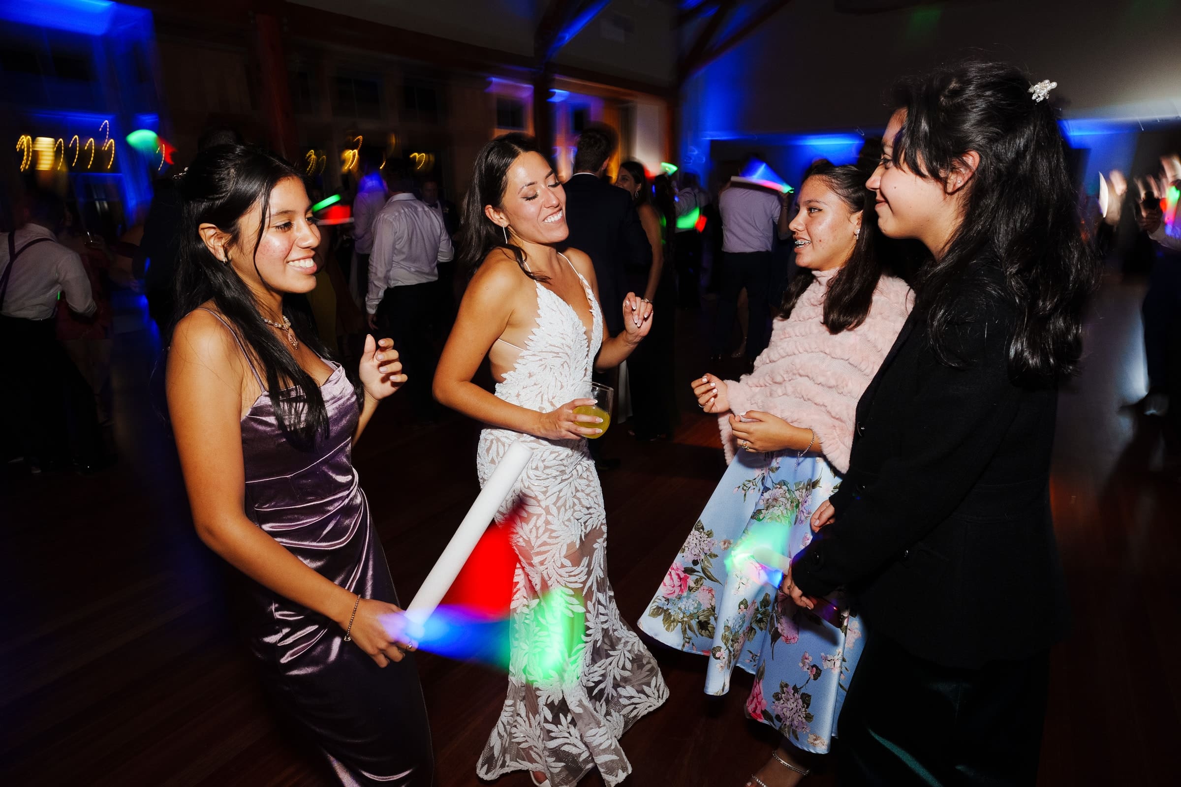 Four women are dancing joyfully at a lively Duke Gardens wedding. Surrounded by colorful lights, two of them wave light sticks with glee. The entire scene is brimming with happiness and festive energy.