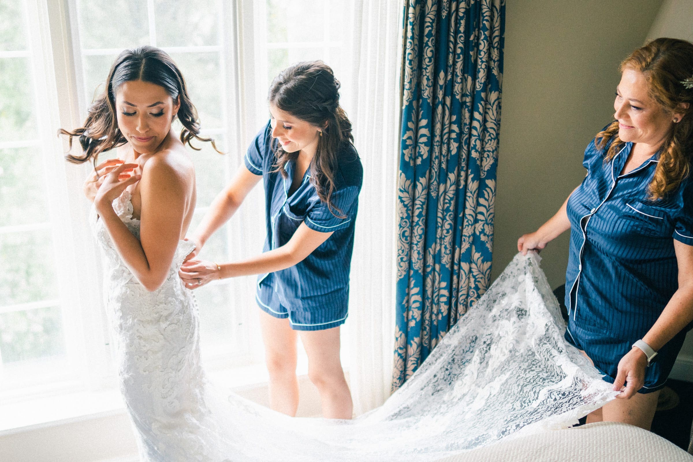 A bride in a lace wedding dress stands near a window at Duke Gardens, while two women in matching navy pajamas assist her. One adjusts the dress, and the other holds the train. Blue patterned curtains flank the window, letting in natural light.