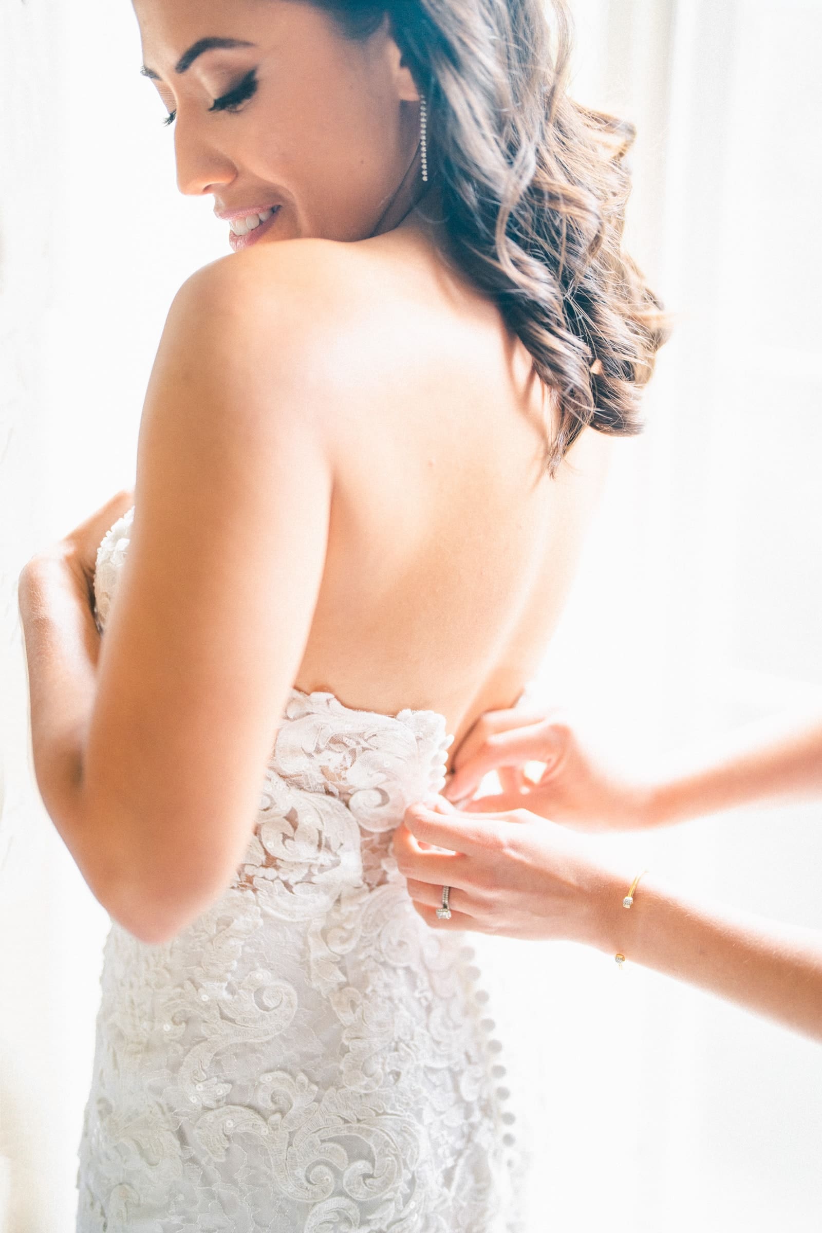 A bride in an elegant white lace dress is being assisted with the back zipper by an unseen person. She smiles softly, with sunlight streaming through a Duke Gardens wedding window behind her, illuminating the intricate floral patterns on her dress.