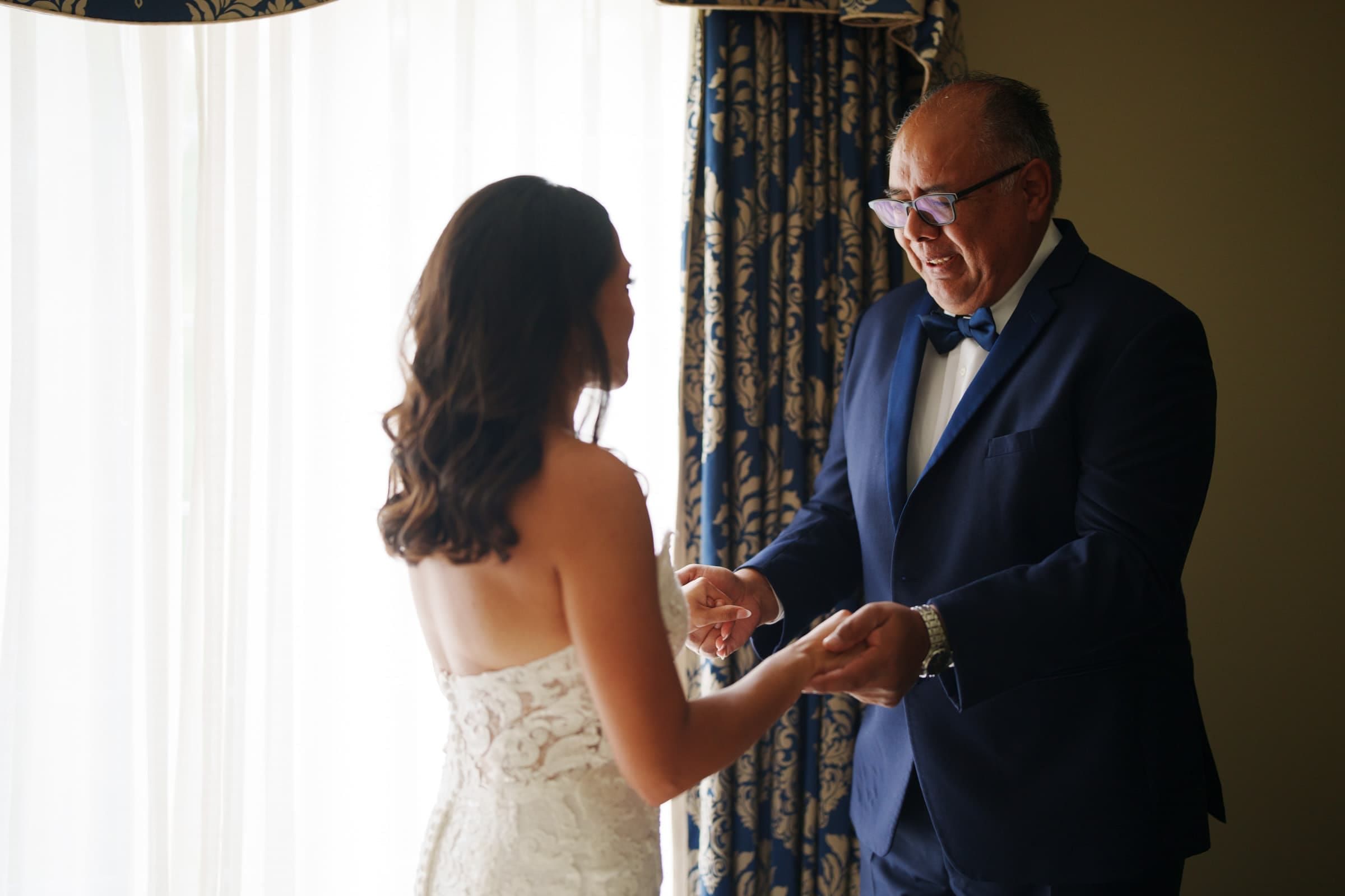 A bride in a white gown shares a joyful moment with a man in a navy suit and bow tie. Together, they hold hands near the window with patterned curtains at their Duke Gardens wedding, both smiling lovingly at each other.