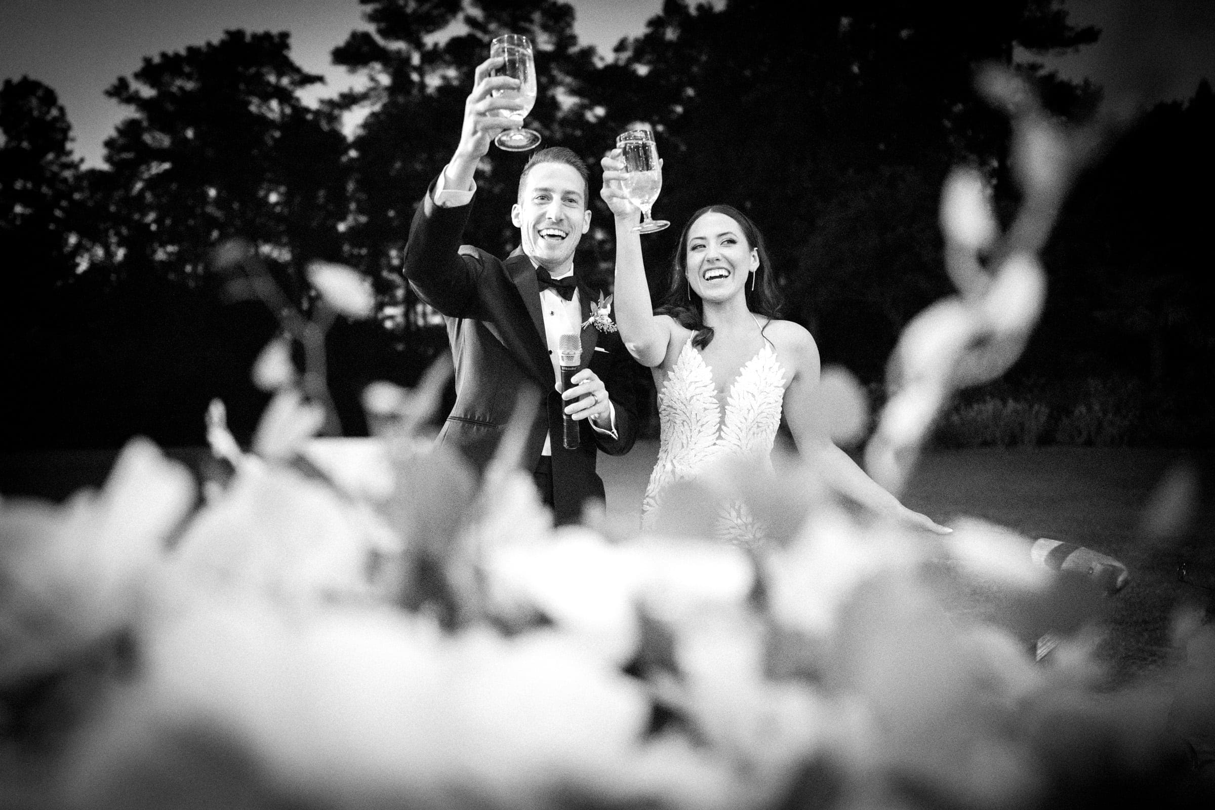 A black and white image captures a smiling couple in formal attire at their Duke Gardens wedding, raising their glasses in a toast. A blurred floral arrangement graces the foreground, with trees softly framing the joyous moment.