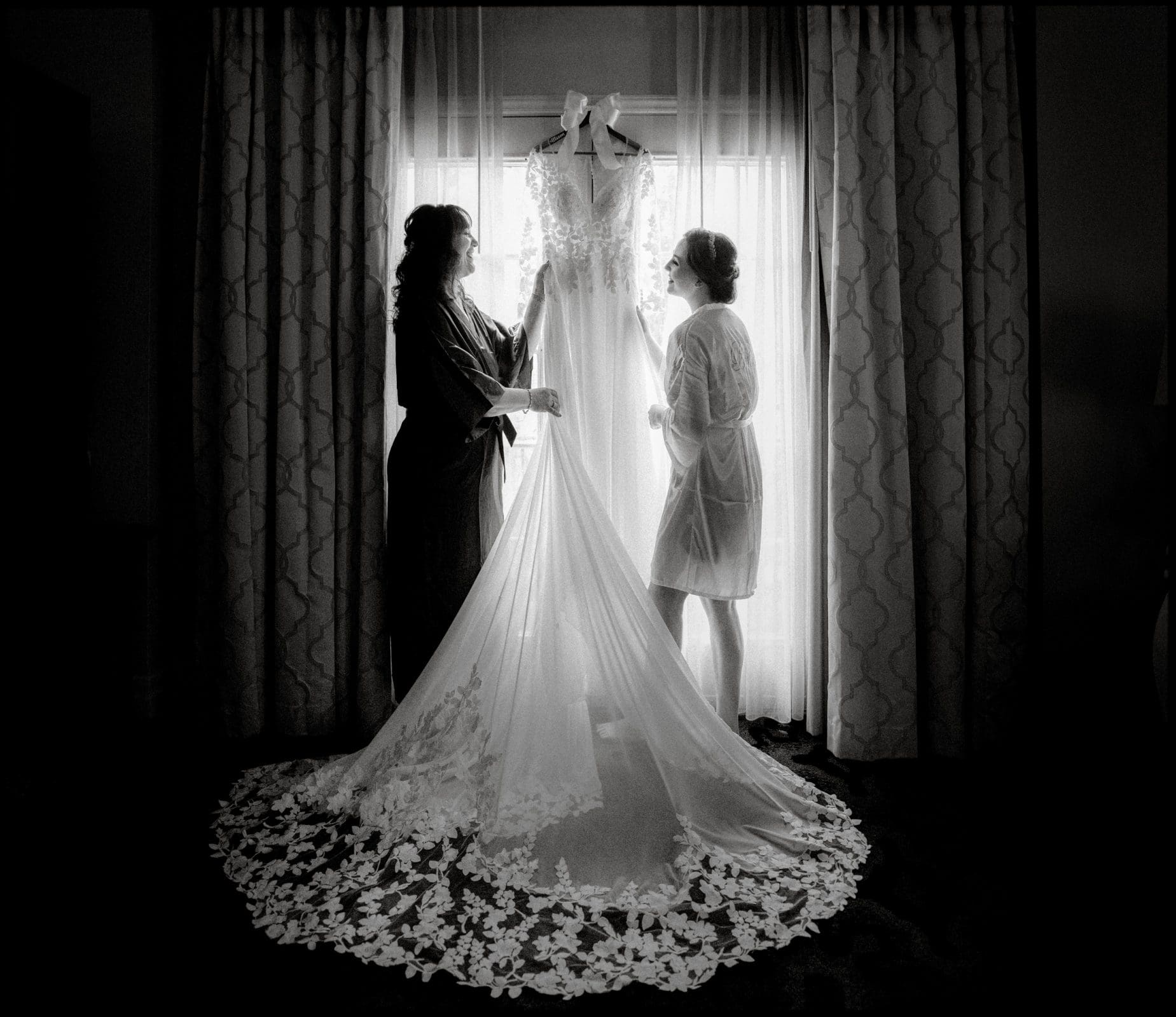 Two women in robes stand before a bright window, admiring a long, lace-trimmed wedding dress hanging from a curtain rod at the Duke Chapel wedding. The dress's train cascades across the floor, forming an elegant silhouette.