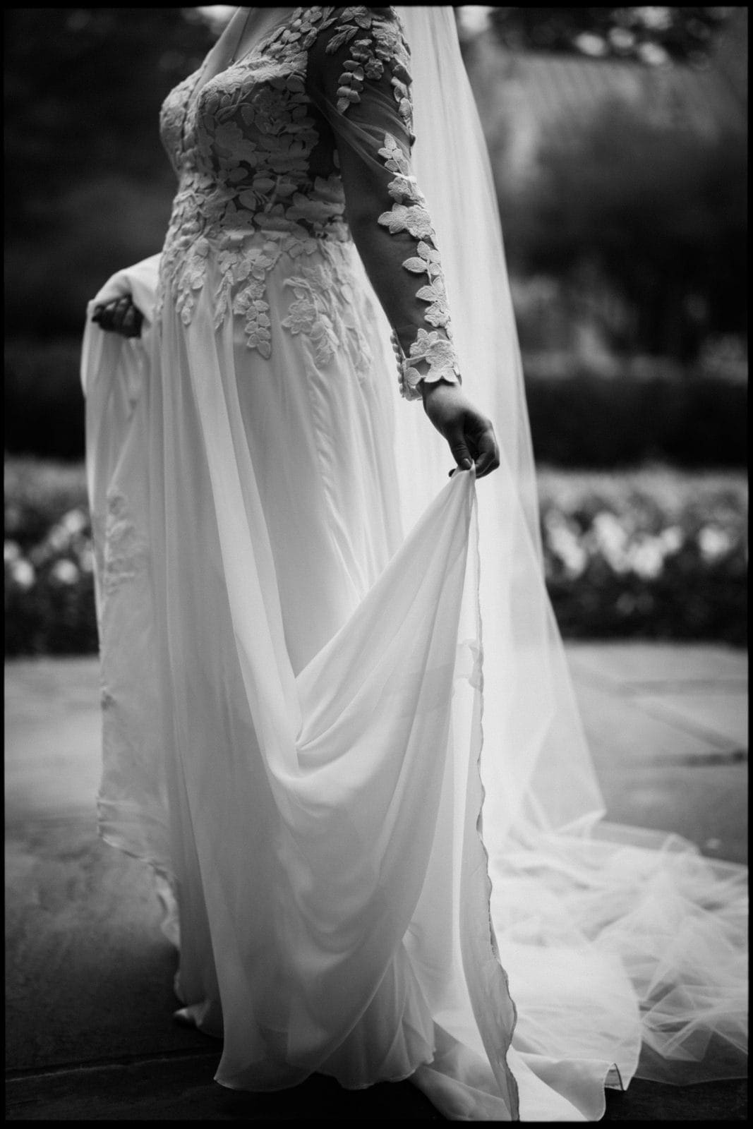 A black and white photo of a bride in a lace wedding dress, holding the skirt delicately. Her veil cascades down her back as she stands on a stone path at Duke Chapel, surrounded by blurred foliage, capturing an air of timeless elegance.
