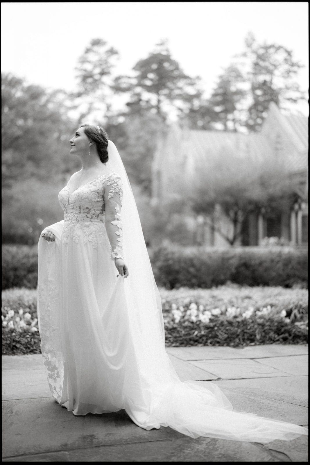 A bride in a long-sleeved wedding gown with lace details stands outdoors at a Duke Chapel wedding, holding her train. She gazes into the distance, surrounded by lush greenery and blurred architectural details. The captivating image is in black and white.