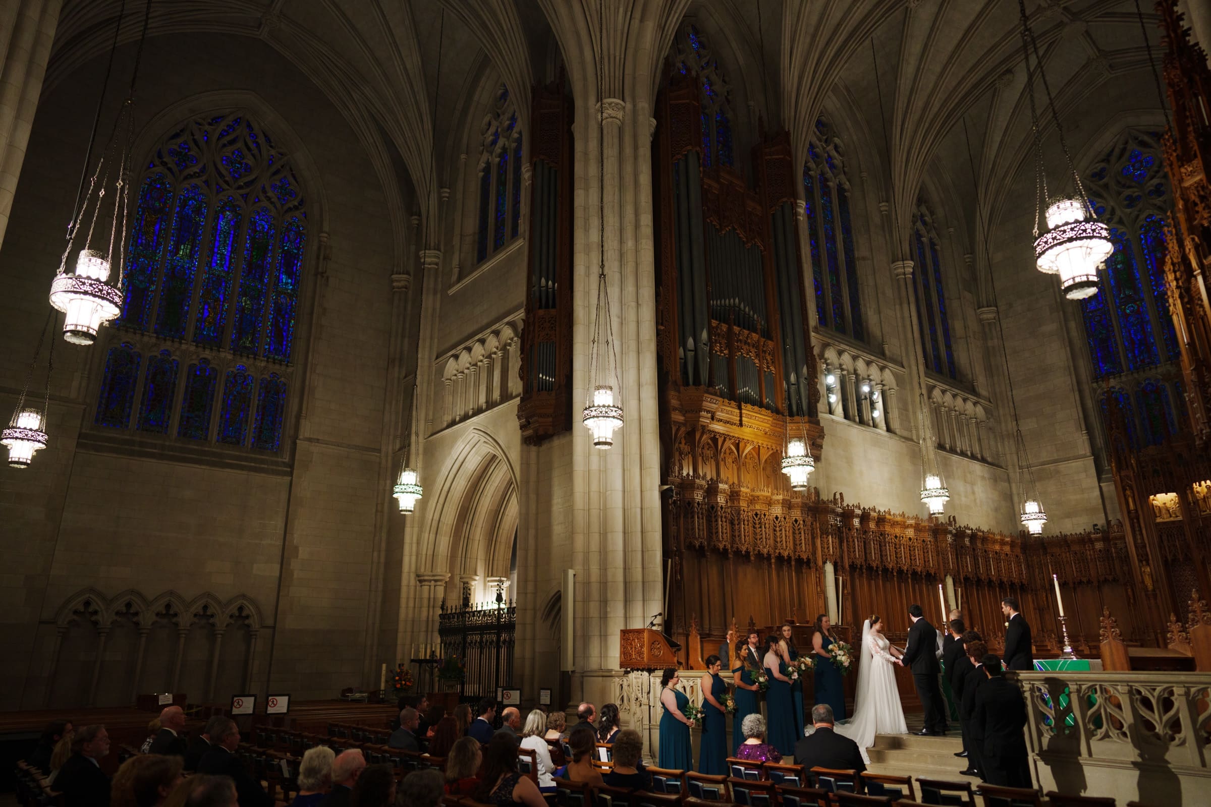 A wedding ceremony unfolds inside the majestic Duke Chapel, a gothic-style sanctuary with high arched ceilings and stunning stained glass windows. The bride and groom stand at the altar, surrounded by their bridal party, as guests are seated in awe of the sacred event.