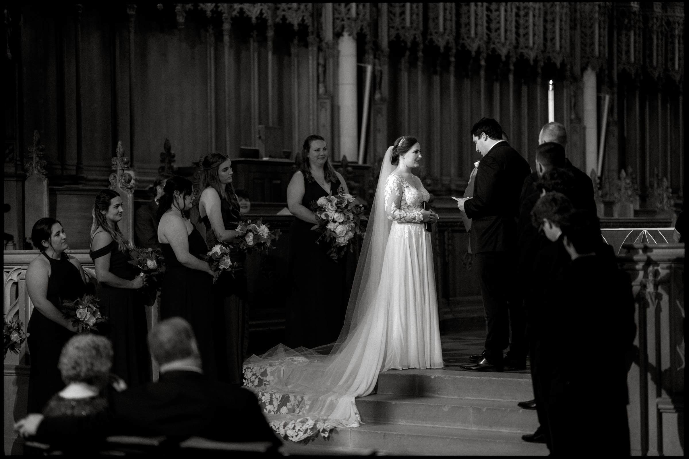 A bride and groom stand on the steps of Duke Chapel, surrounded by a bridal party. The bride is in a long gown with a veil, and the groom is in a suit. Bridesmaids hold bouquets. The scene is solemn and elegant, with the intricate details of the chapel adding to its timeless beauty.