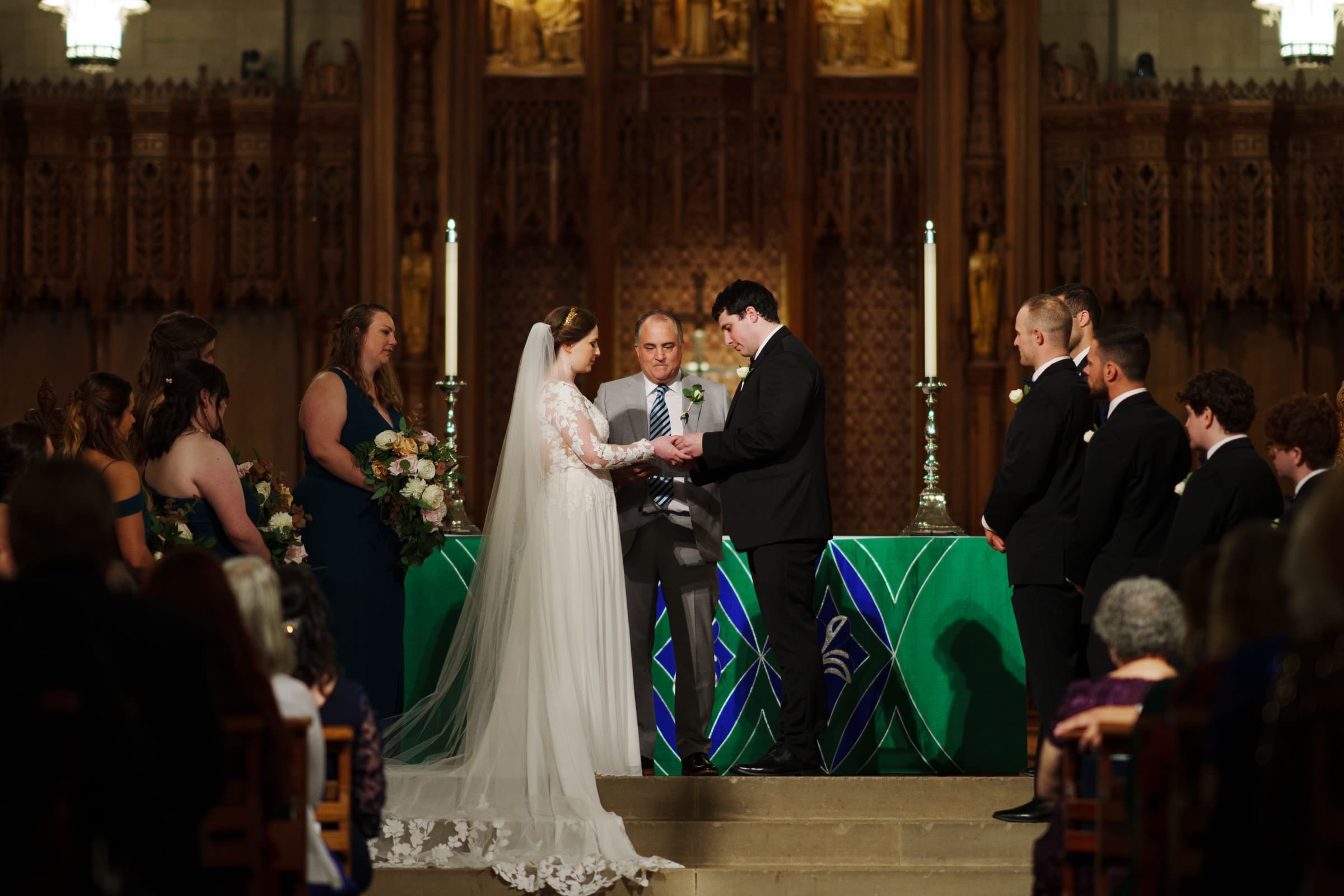 At the altar of Duke Chapel, a bride and groom exchange rings amid a breathtaking wedding ceremony. The bride dazzles in her white dress and veil, while the groom stands poised in a black suit. Bridesmaids and groomsmen flank them as guests watch intently from their seats.