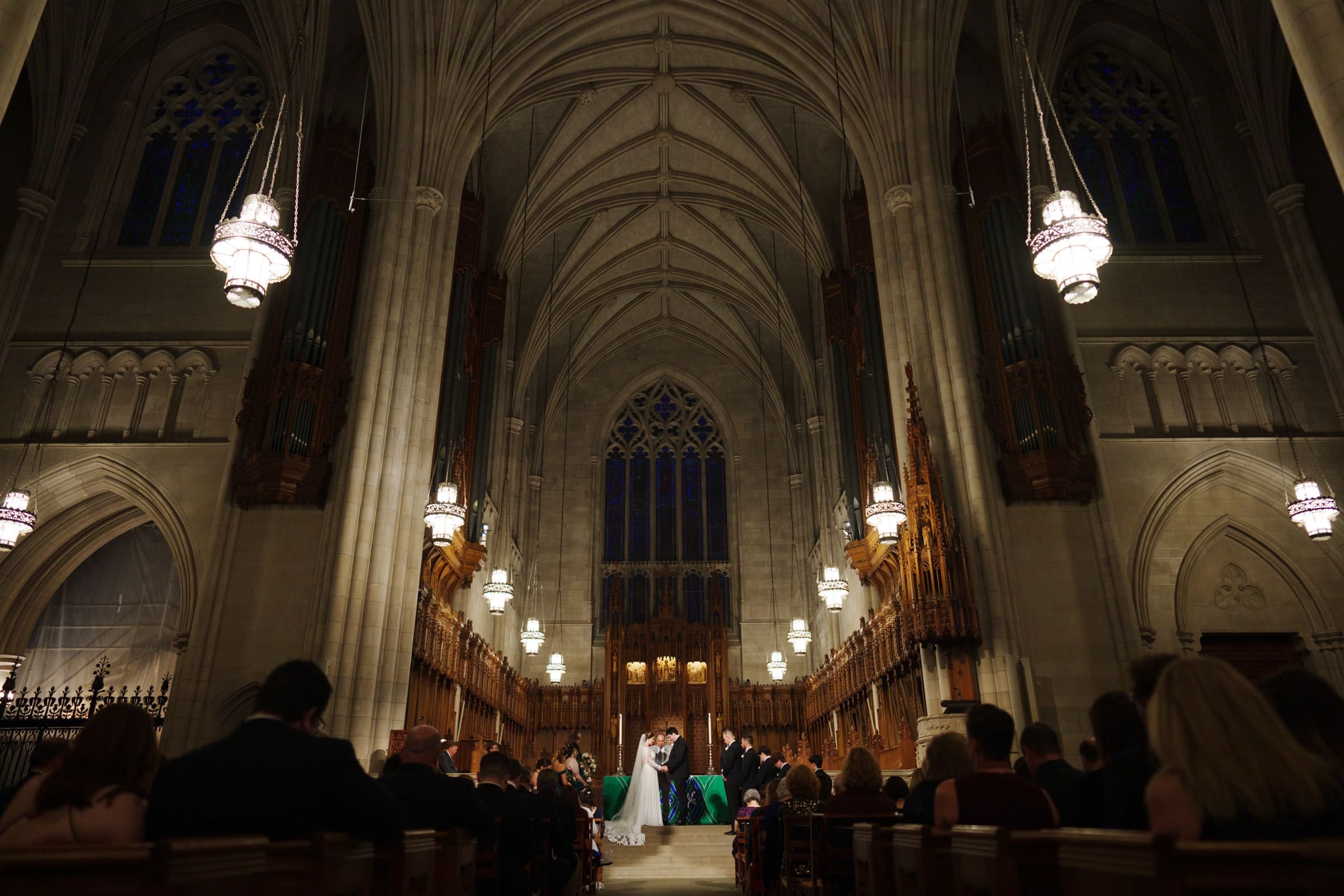 A couple stands at the altar in Duke Chapel, a grand cathedral with vaulted ceilings and ornate chandeliers. Guests are seated in pews, witnessing the ceremony. Gothic architecture and dim lighting create a solemn and majestic atmosphere.