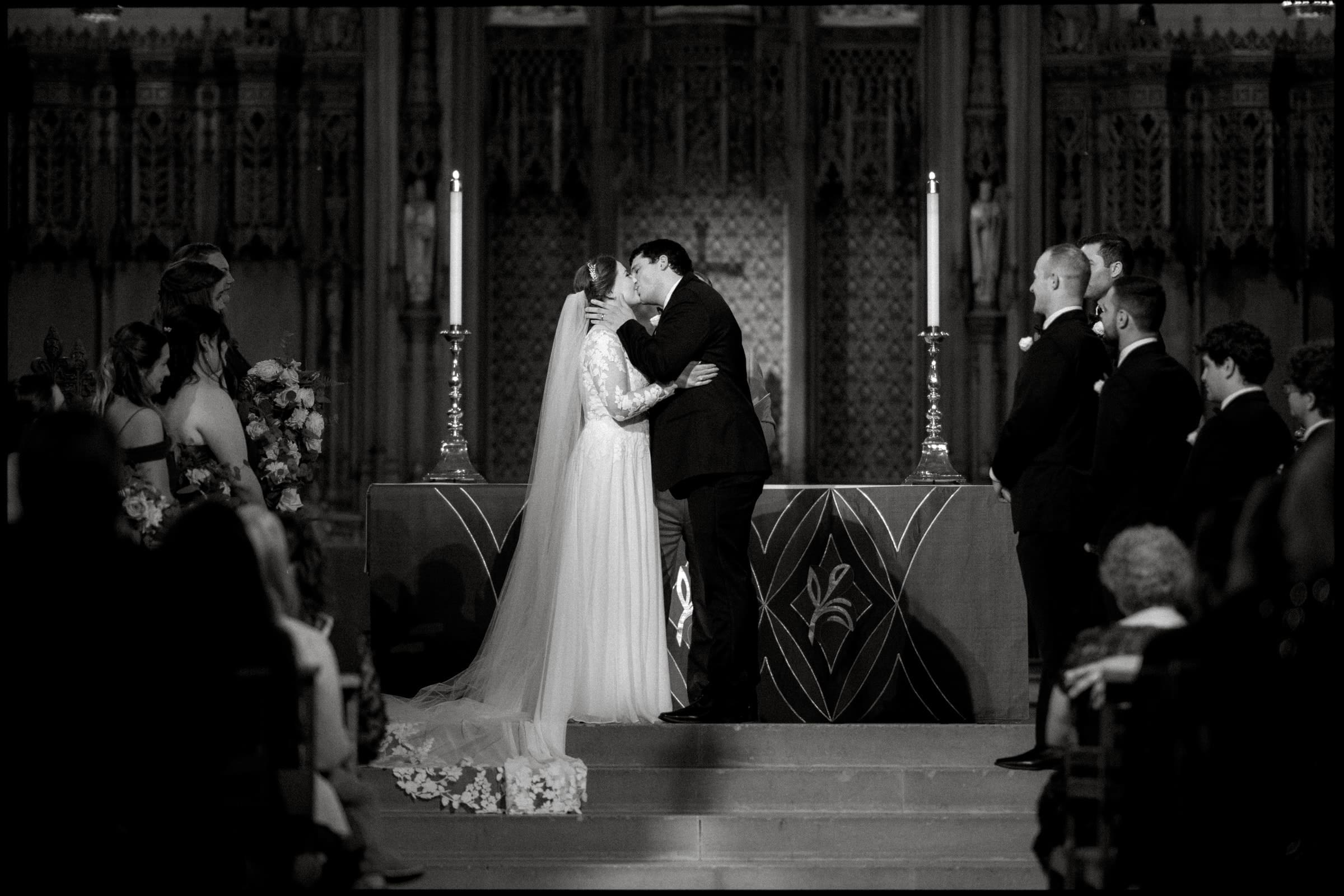 In a timeless black-and-white photo of a Duke Chapel wedding, a couple shares a tender kiss at the altar. The bride, in her gown and long veil, and the groom, in a sharp suit, are surrounded by their joyous wedding party. Candles flicker amid the chapel's ornate details in the background.