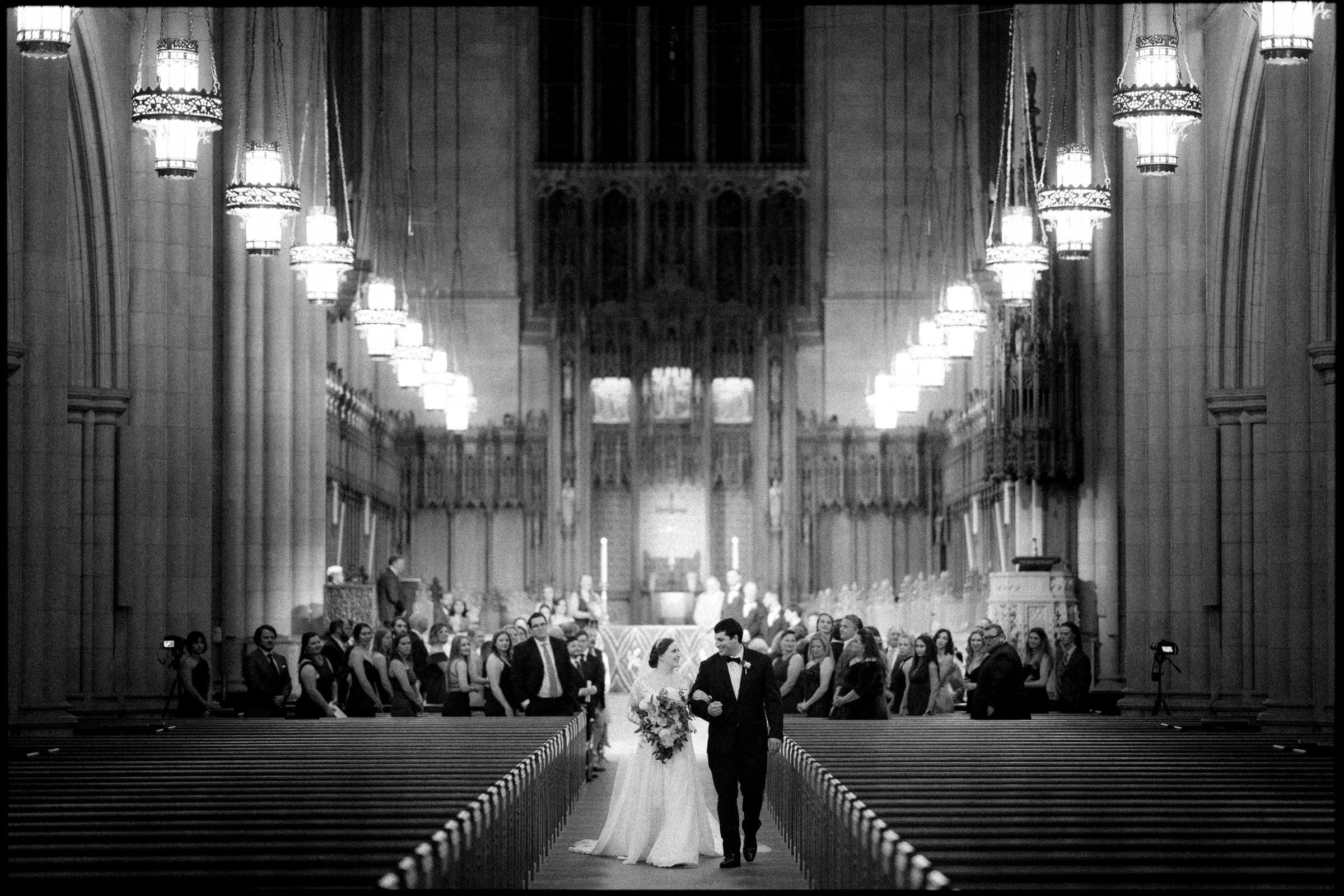A black and white photo captures a bride and groom walking down the aisle of the grand Duke Chapel, its glowing chandeliers illuminating their path. Guests stand on either side, admiring the couple under the high, arching ceiling of this ornate church.