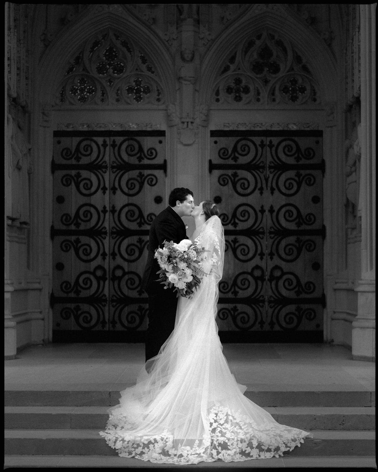A bride and groom share a kiss on the steps of an ornate building, reminiscent of a Duke Chapel wedding. The bride wears a long lace train gown and holds a bouquet. The building features intricate, arched doors with decorative patterns. Black and white photo.