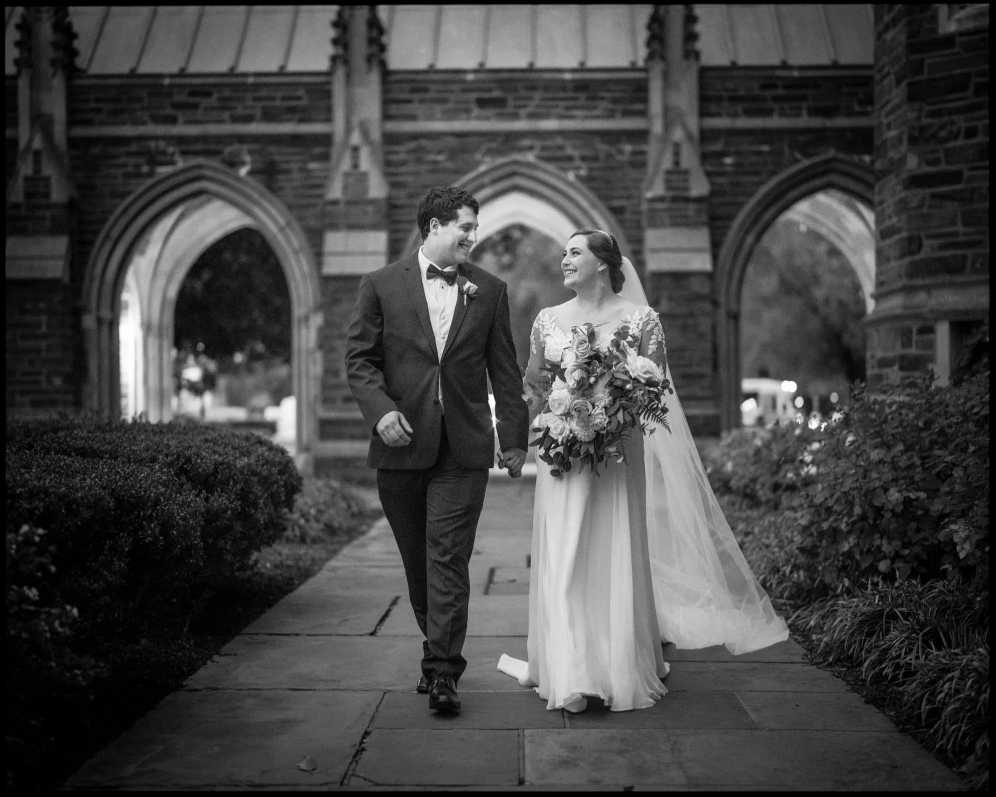 A bride and groom walk hand in hand outside, smiling at each other. The bride holds a bouquet and wears a flowing dress with a long veil. The groom dons a suit with a bow tie. They stroll along the stone path of Duke Chapel, arches standing majestically in the background.