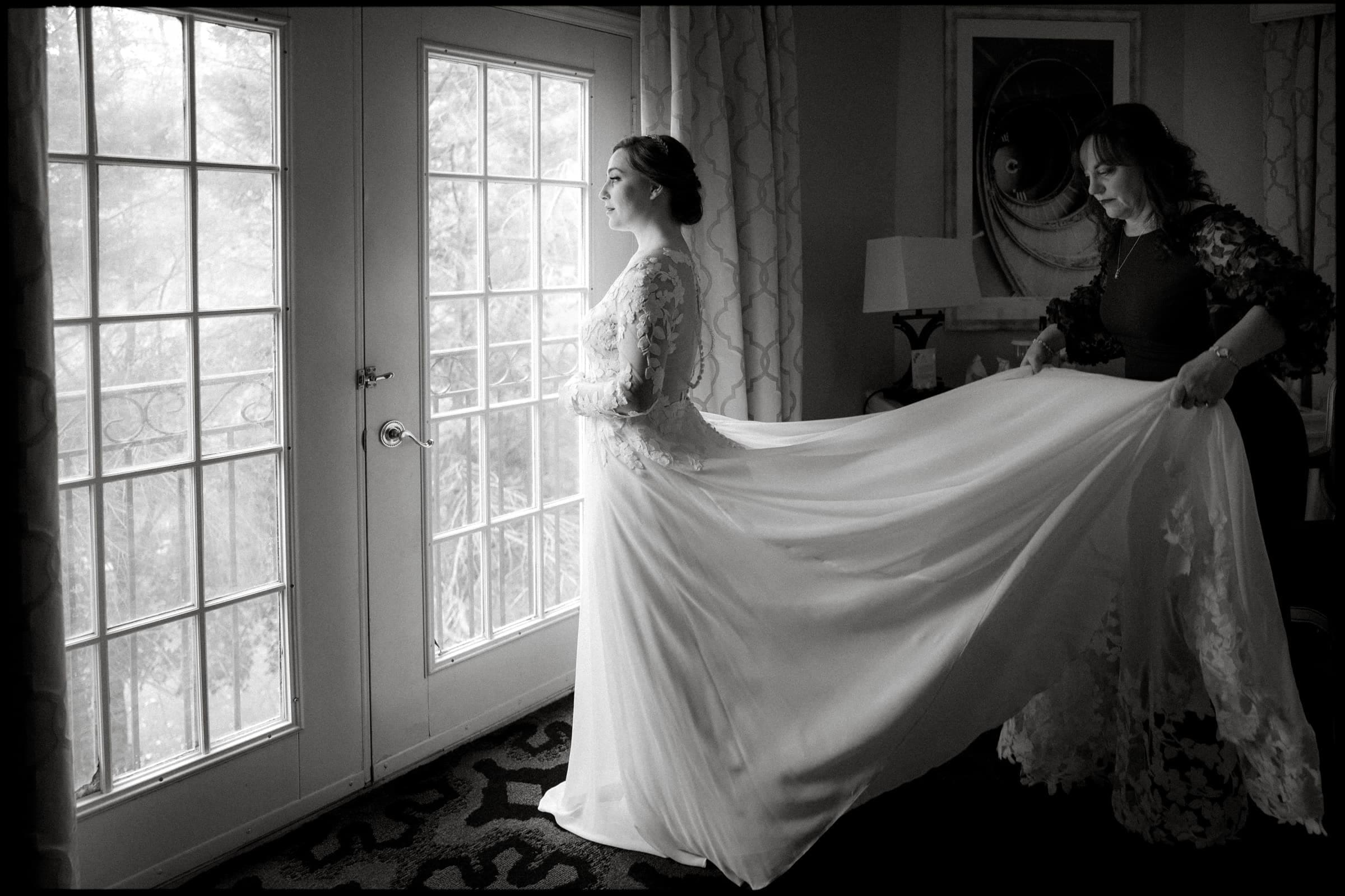 A bride in a long, flowing dress stands by the glass doors of Duke Chapel, staring outwards with a serene expression. A woman assists, holding the train of the dress. The scene is captured in soft, natural light, highlighting the elegance and anticipation of this wedding moment.