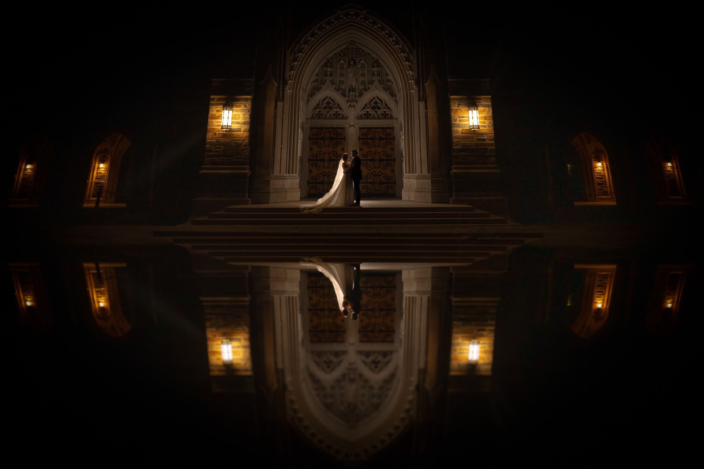 A bride and groom stand on the steps of Duke Chapel, a grand, dimly-lit church with ornate wooden doors. Their reflection shimmers on a glossy surface in the foreground, crafting a symmetrical effect in the shadowy ambiance.