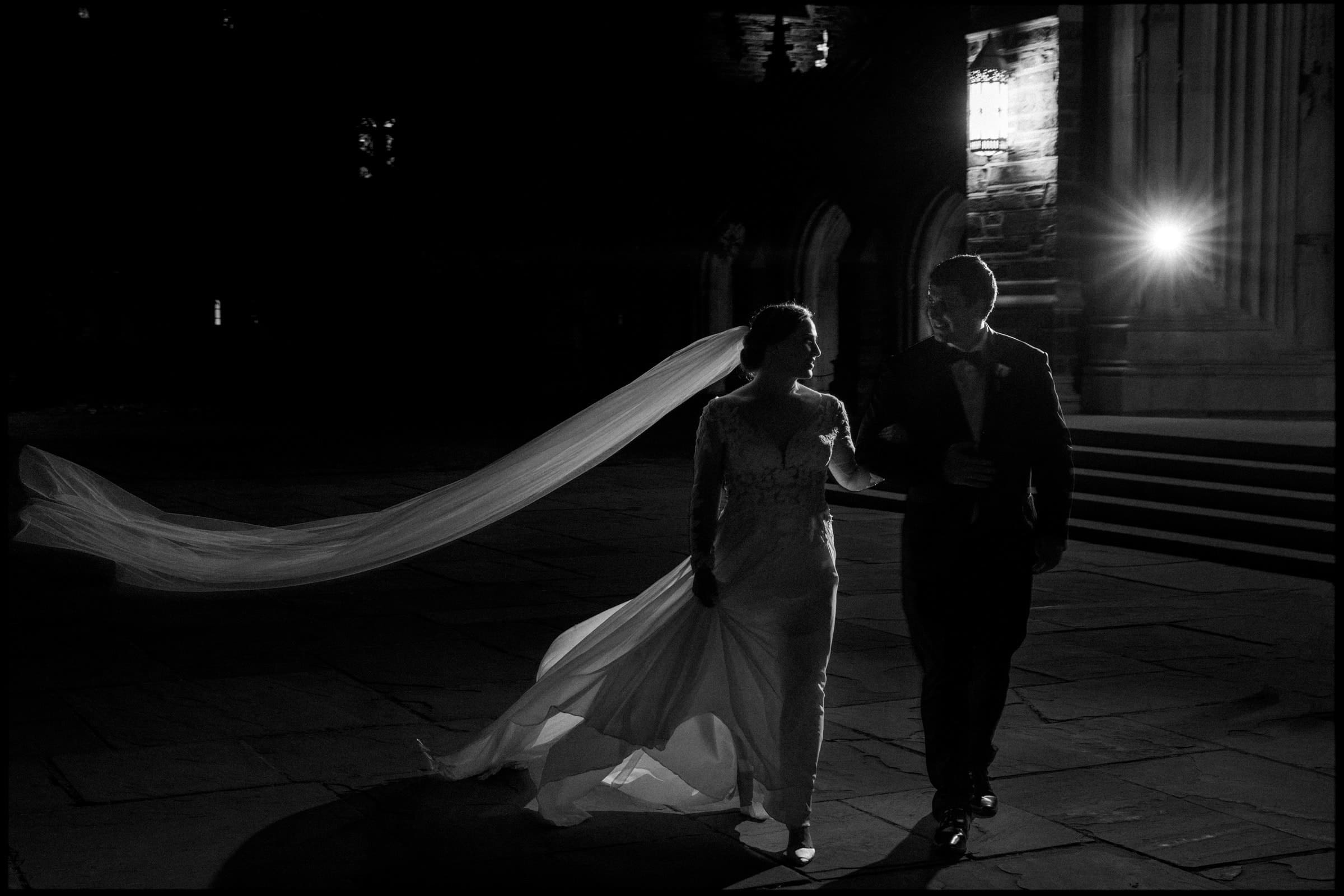 A bride and groom walk together at night, illuminated by a single light. Her long veil flows dramatically in the wind as they stand in front of the grand Duke Chapel with stone steps. The scene is in black and white, adding a classic feel to their enchanting wedding moment.